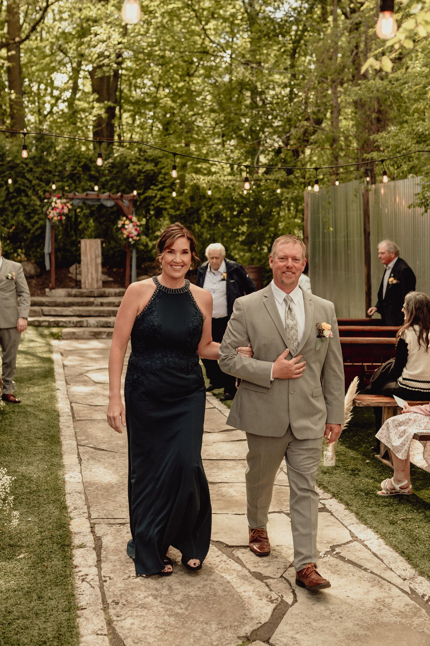 A couple in formal attire walks together on a stone path at an outdoor wedding reception with string lights overhead.
