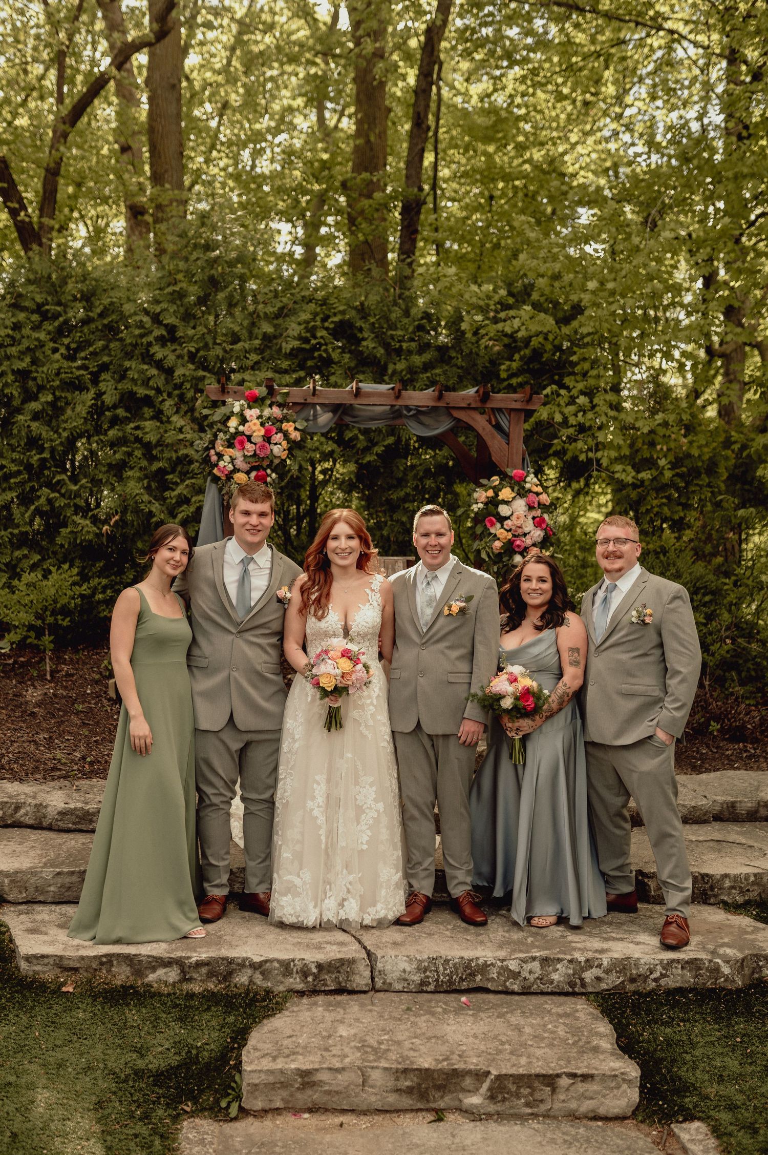 Wedding party in sage green and gray attire poses together on stone steps surrounded by lush greenery.
