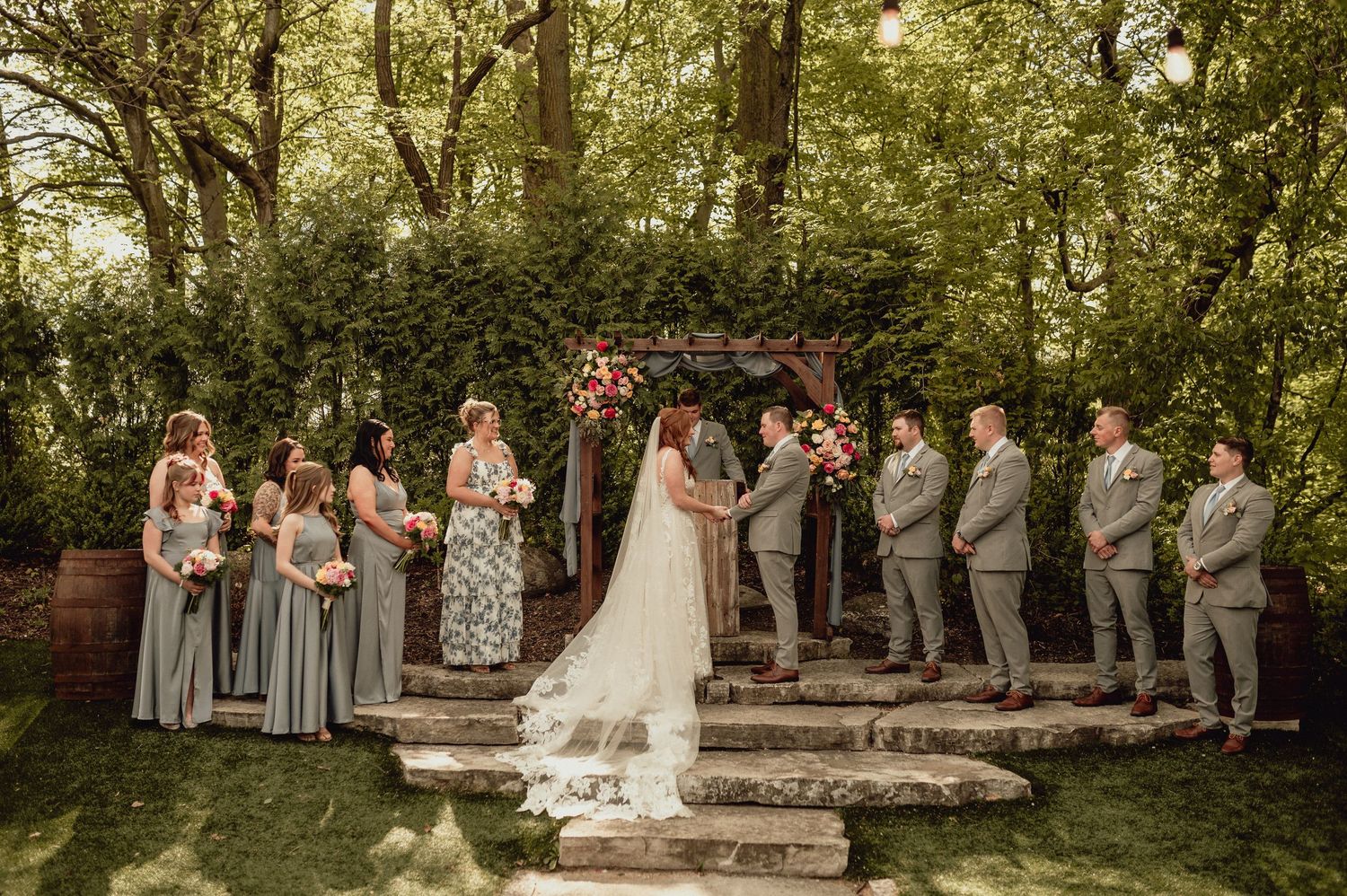 Wedding party poses together in a lush garden setting with bridesmaids in gray dresses and groomsmen in suits.