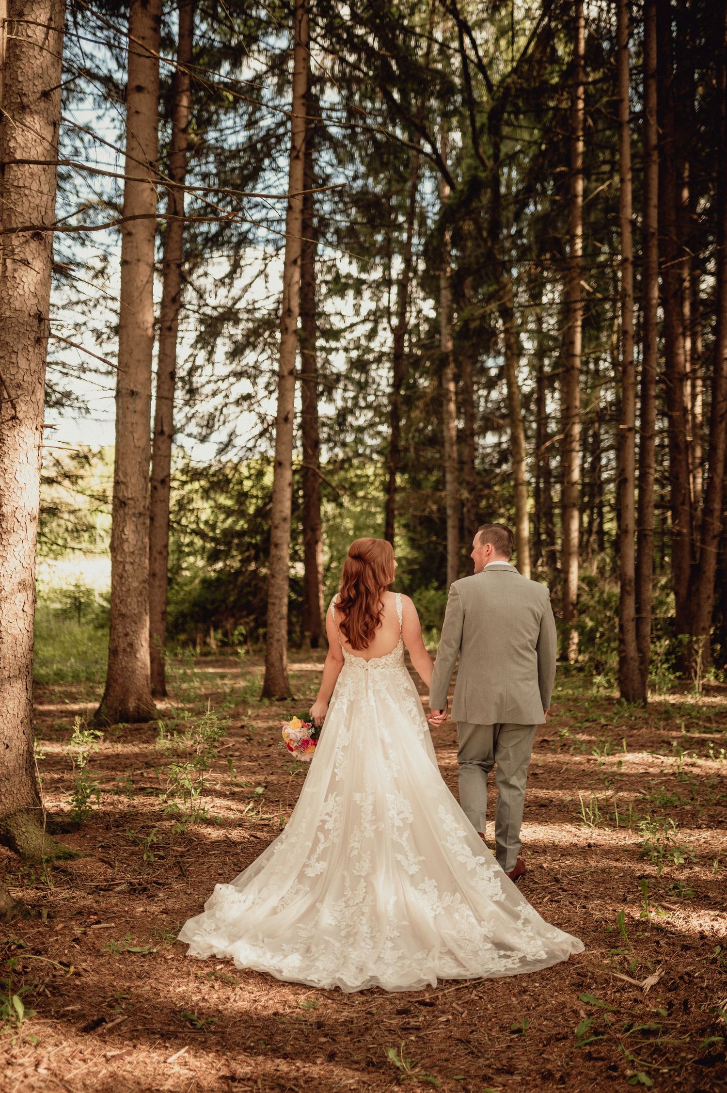 A couple in wedding attire walks through a sunlit pine forest with long shadows cast on the autumn ground.