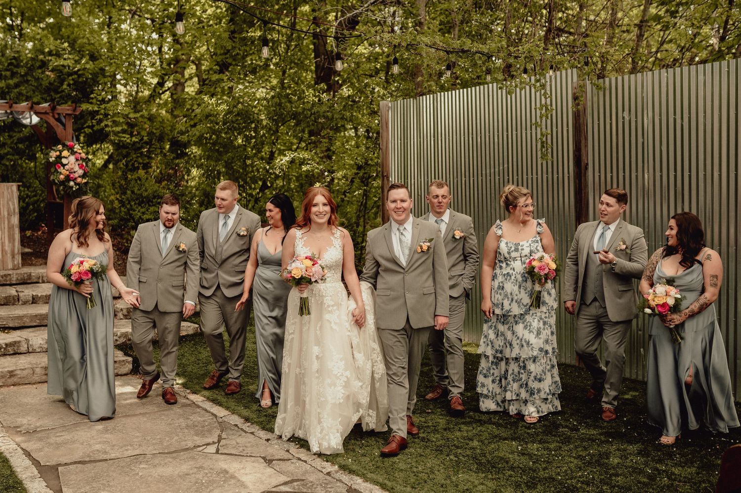 Wedding party in sage green and gray attire poses outdoors against a garden backdrop during summer celebration.