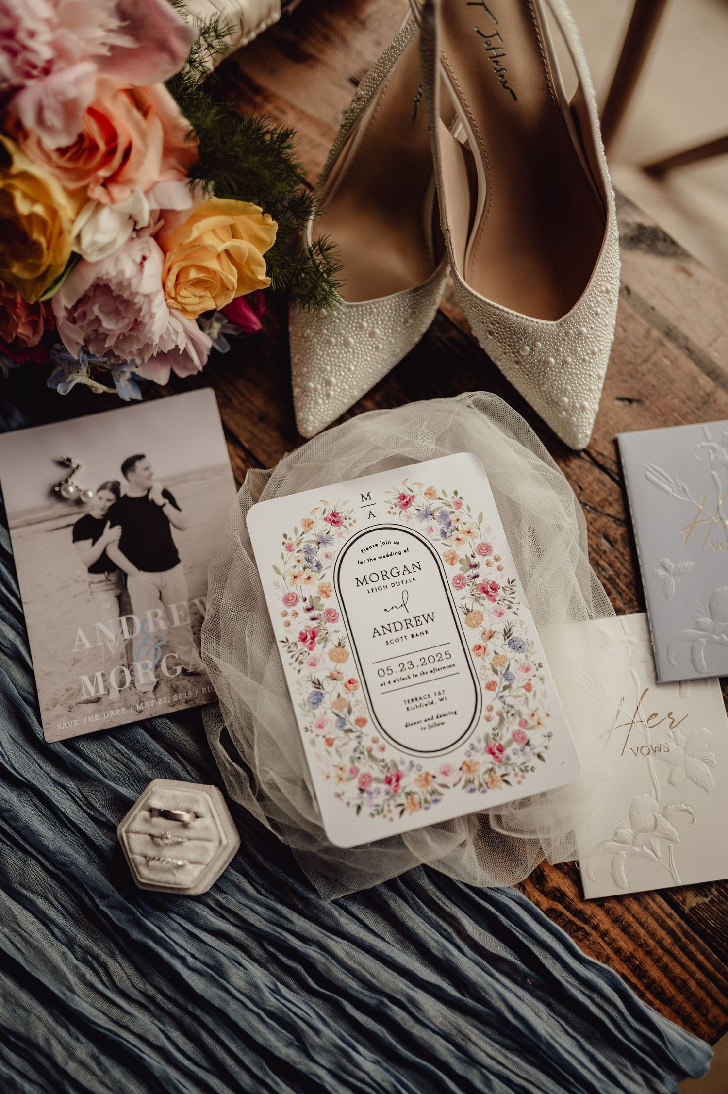 Elegant wedding details including floral invitations, perfume bottles, and delicate accessories arranged on textured fabric.