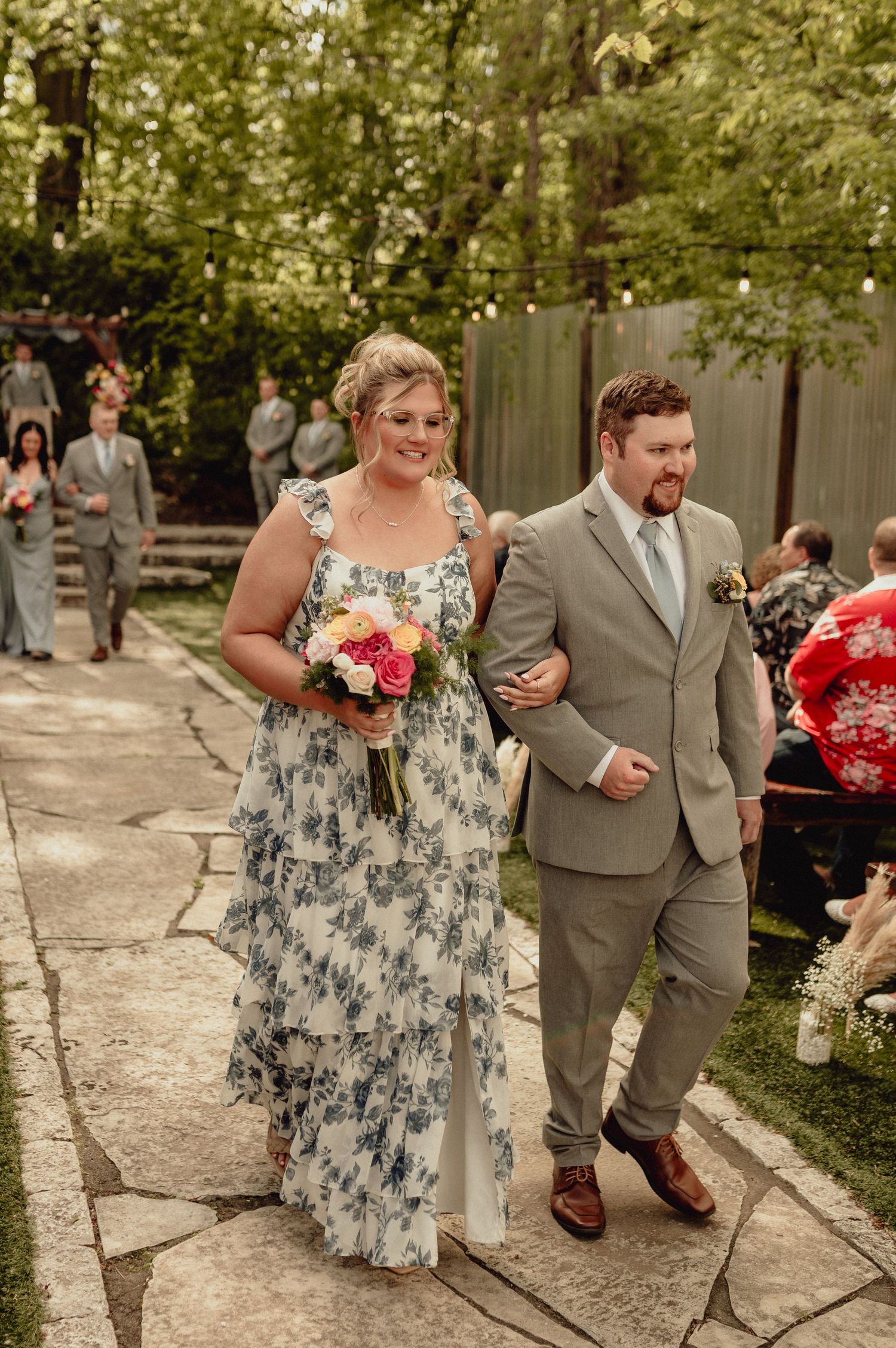 A couple in a floral dress and gray suit walk together on a stone path during an outdoor wedding celebration.
