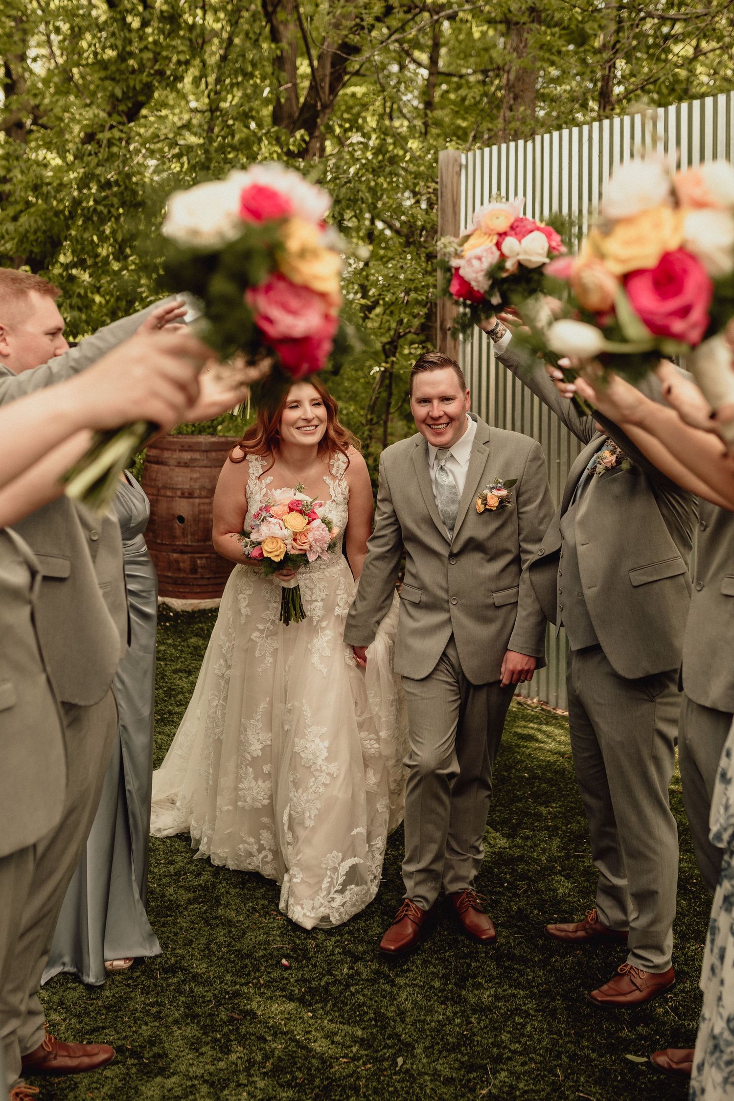 Wedding party in gray suits and champagne dress pose playfully with pink bouquets in garden setting.