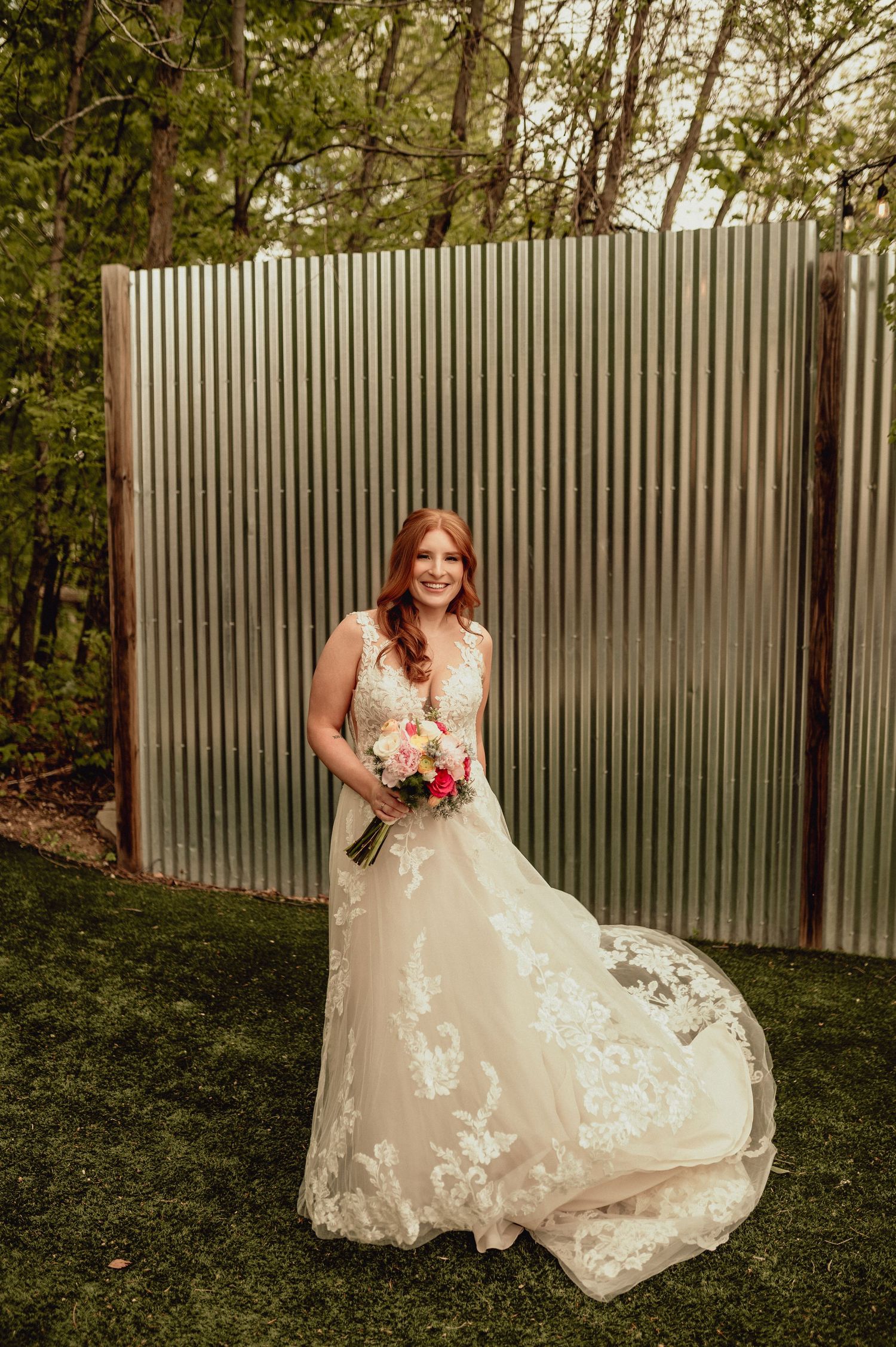 A bride in a lace wedding dress poses outdoors against a corrugated metal fence in a garden setting.