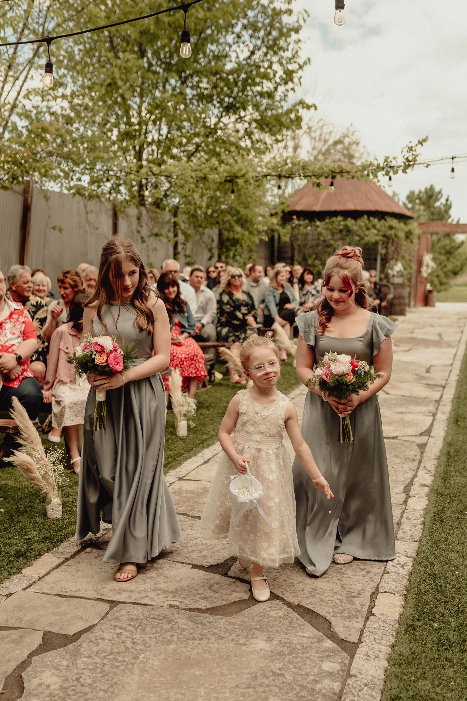 Wedding party in gray dresses and coral bouquets poses playfully on a stone path outdoors during spring celebration.