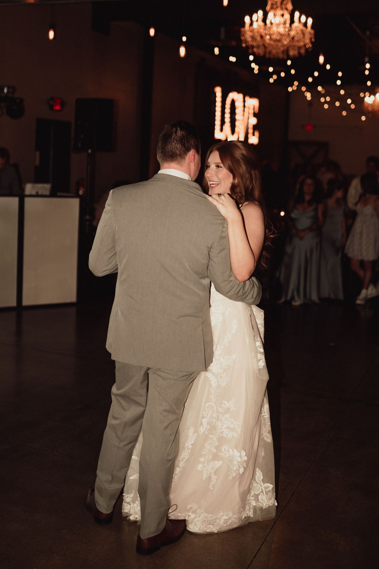 A couple dances together at a wedding reception under a crystal chandelier and twinkling lights.