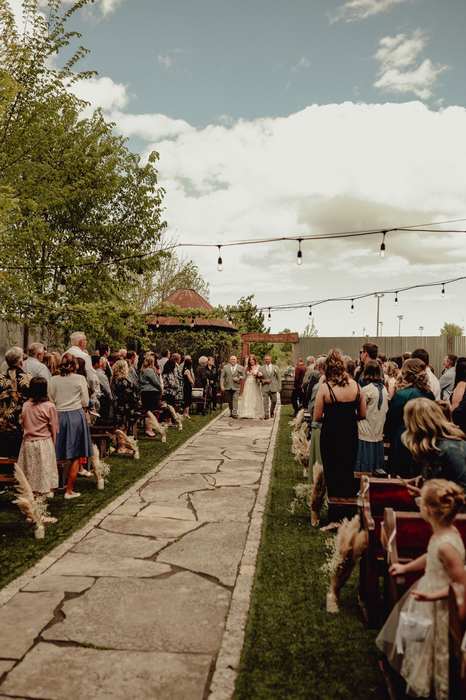 Wedding guests line a stone pathway under string lights during an outdoor ceremony at sunset.