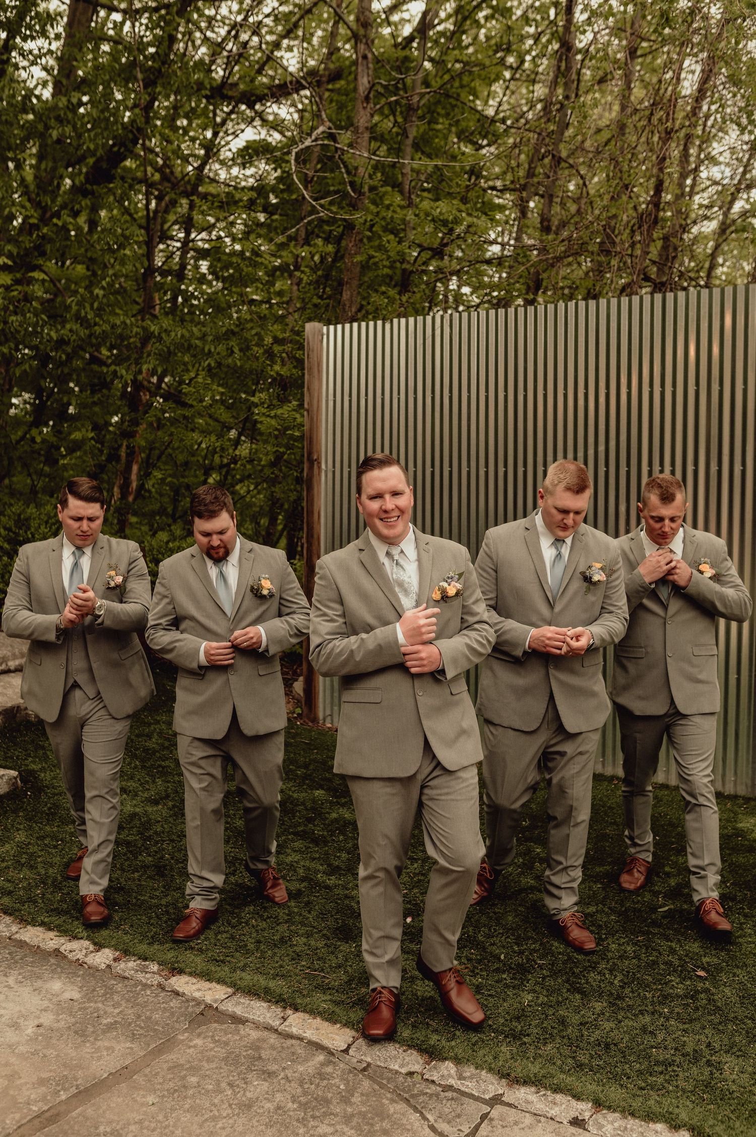 A wedding party in gray suits stands in a line against a hedge while looking at their phones.