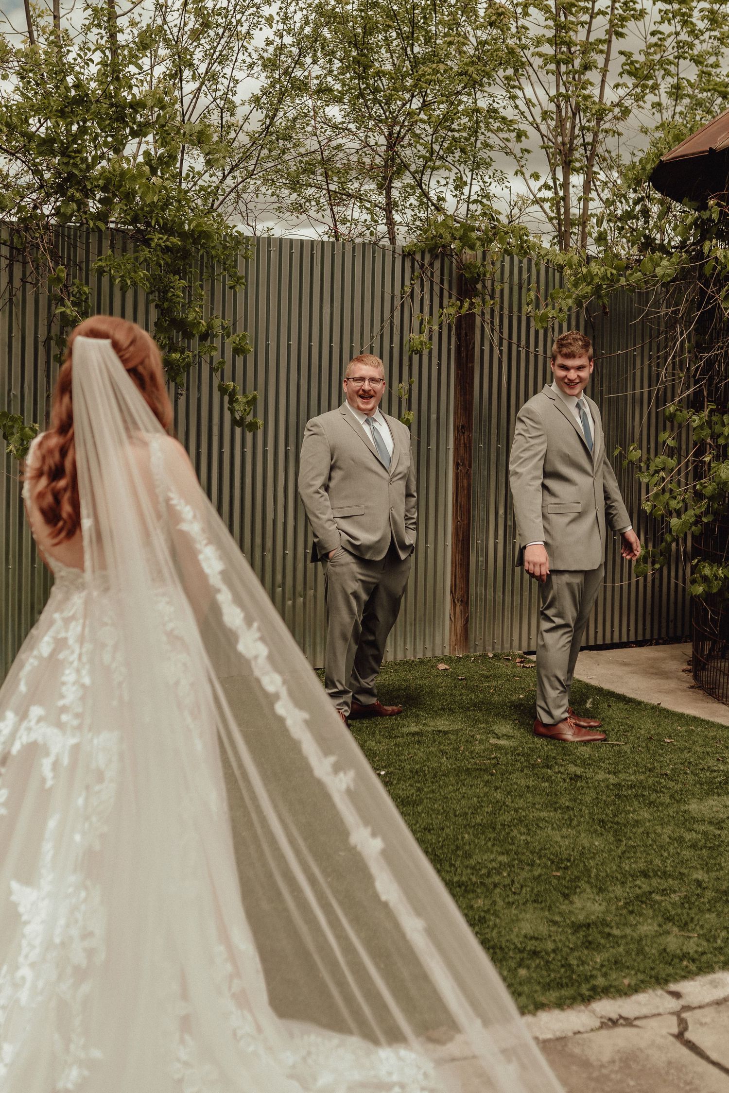 A wedding first look moment captured in a garden setting with a flowing white veil and light gray suit.