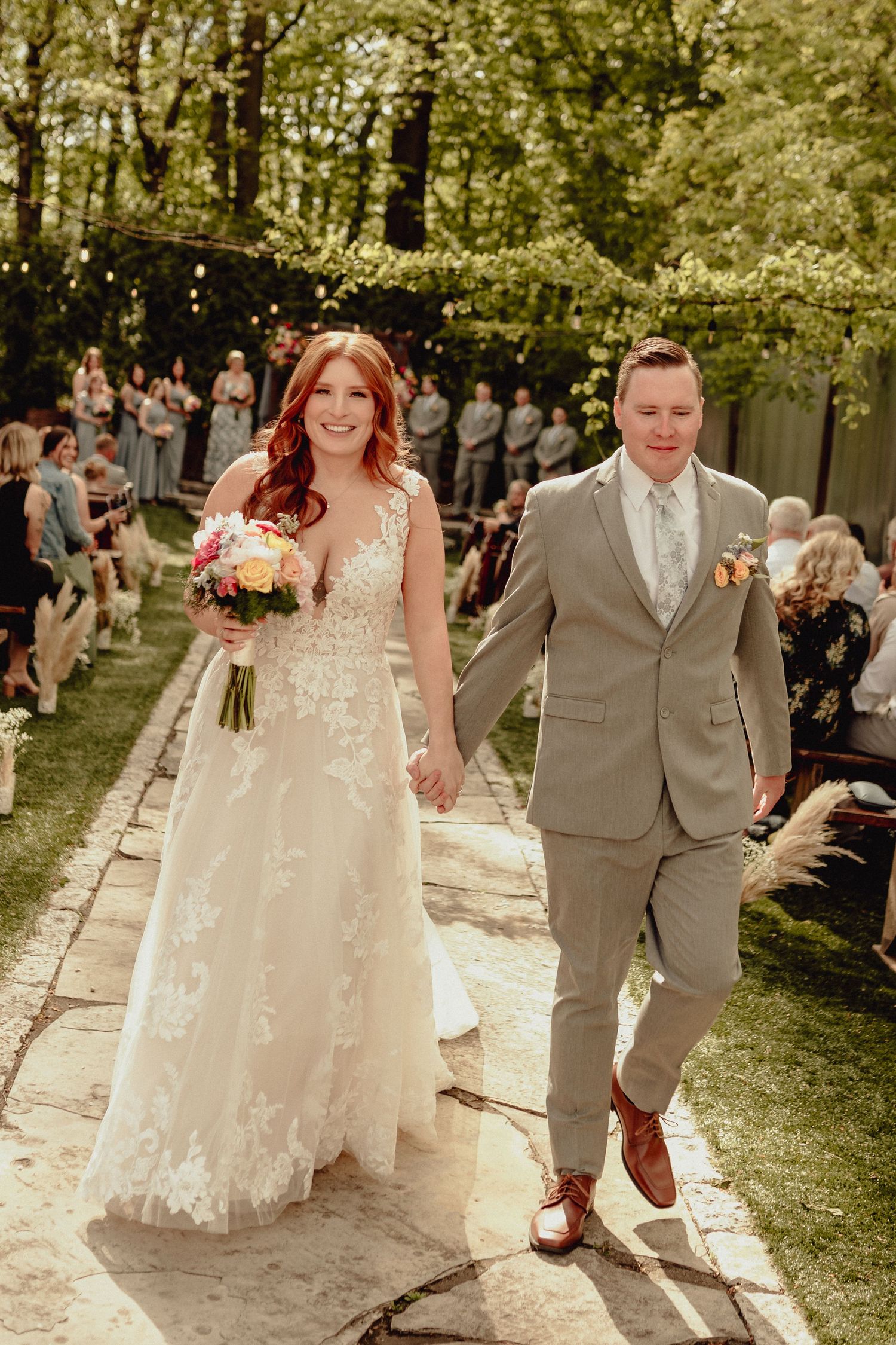 A wedding couple walks joyfully down a sunlit garden path while guests look on during an outdoor ceremony.