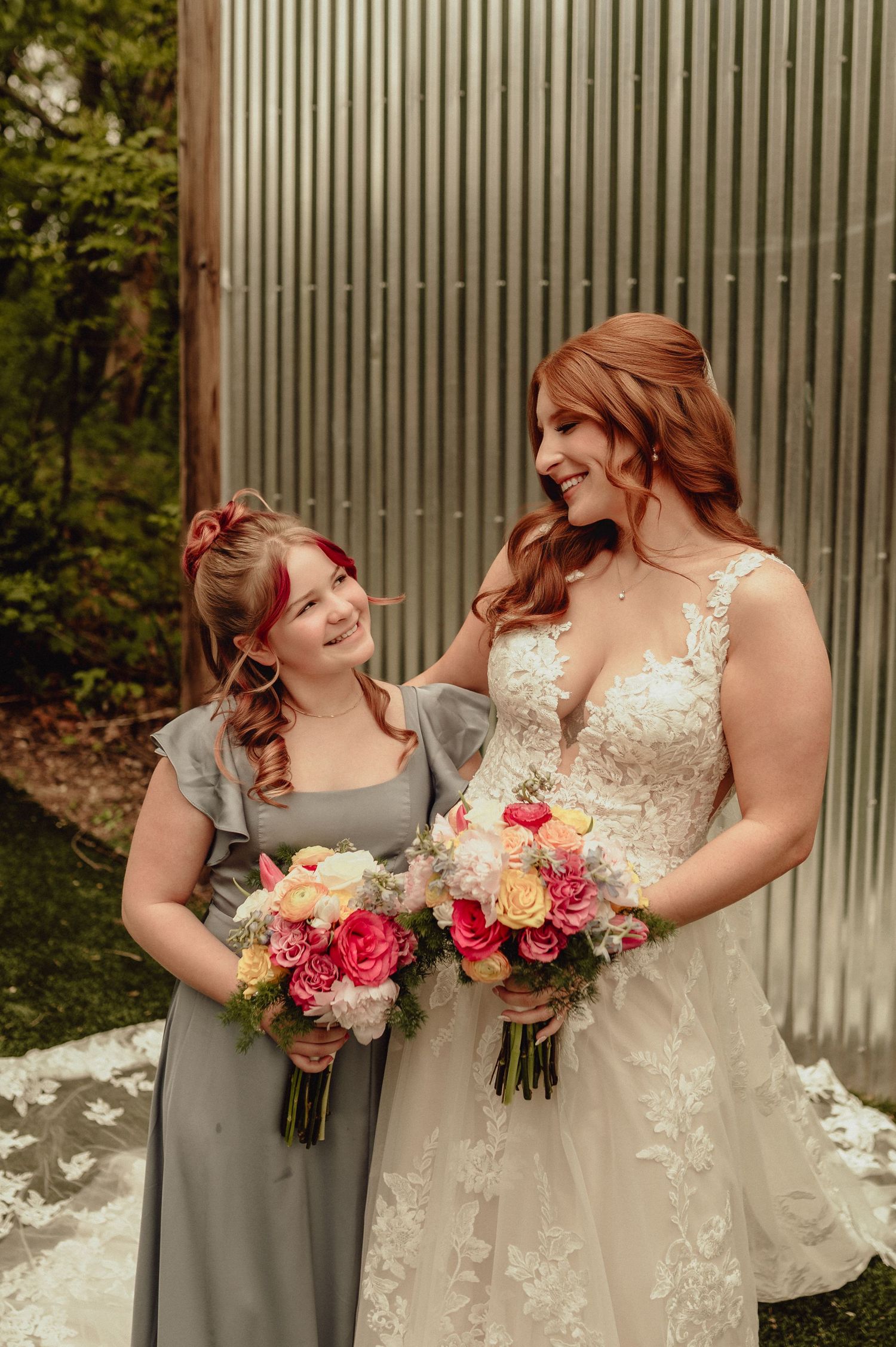Two people in wedding attire share a joyful moment while holding vibrant bouquets of roses and flowers against a metal wall.