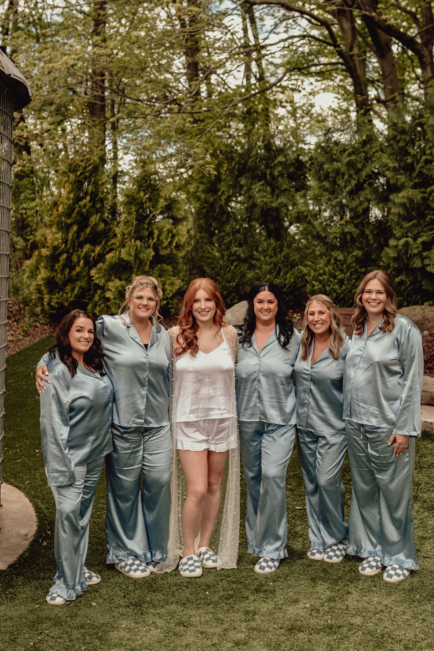 A wedding party in matching blue silk robes poses playfully on grass near trees during a celebration.
