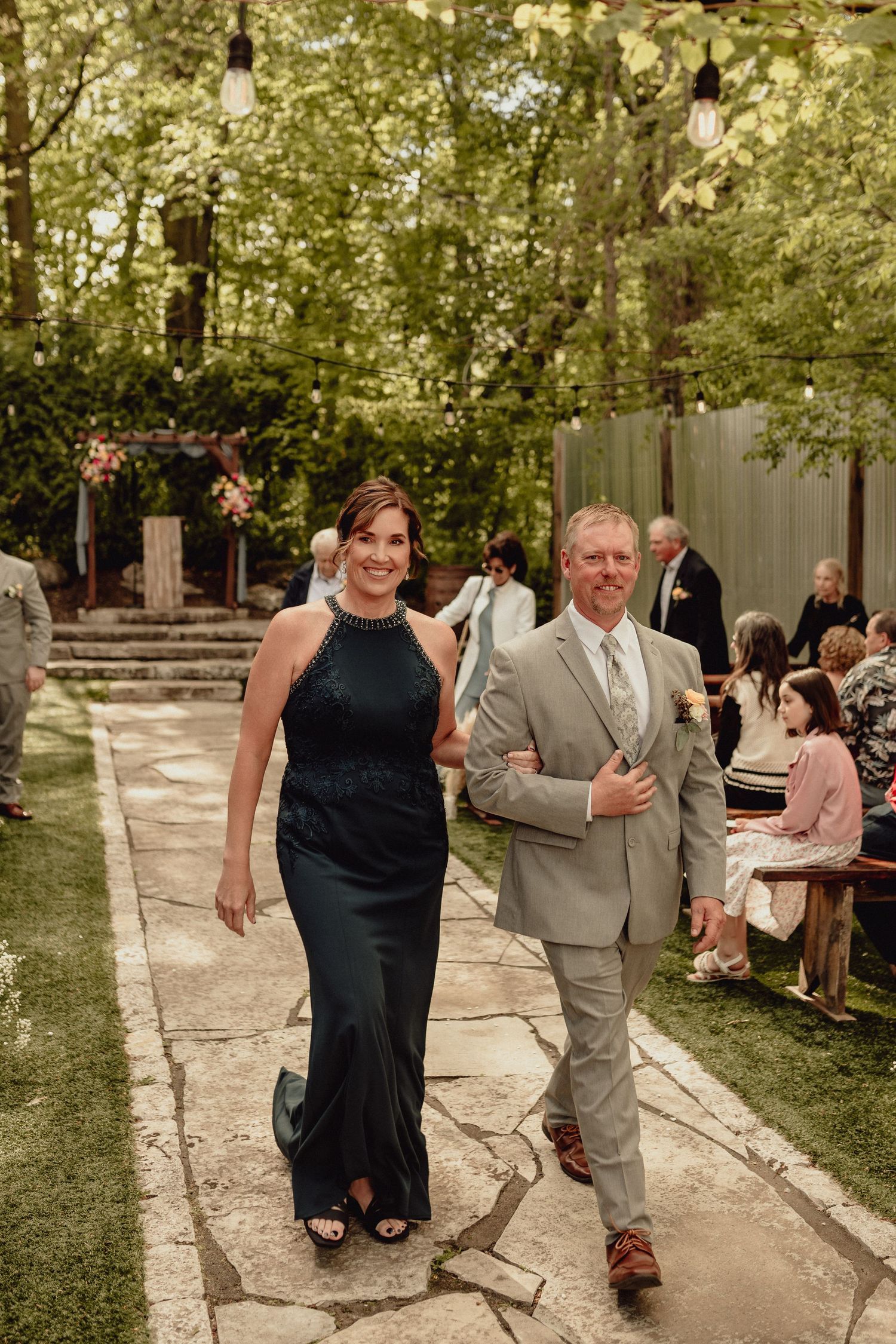 Wedding guests walk along a stone path through a garden setting during an outdoor celebration.