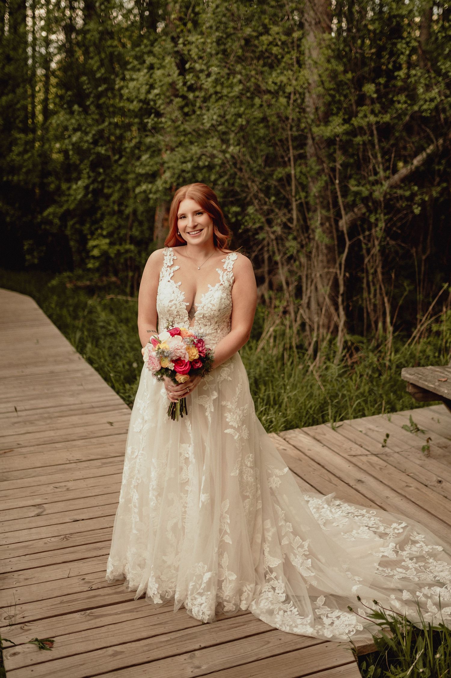 A bride in a lace wedding gown with a plunging neckline and flowing train stands on a wooden boardwalk surrounded by greenery.