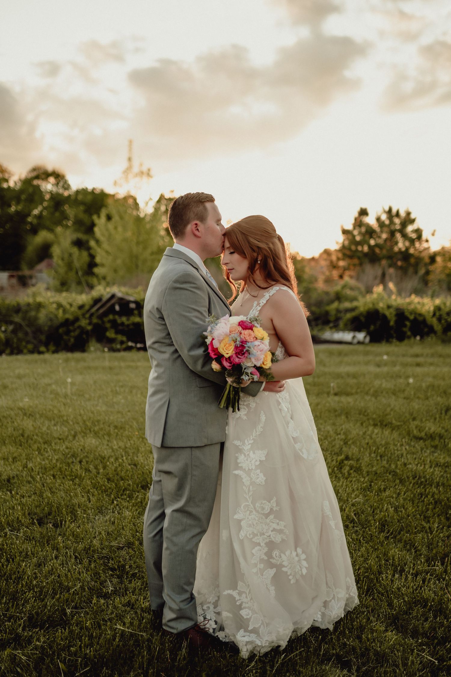 A romantic wedding couple shares an intimate moment in a grassy field at sunset.