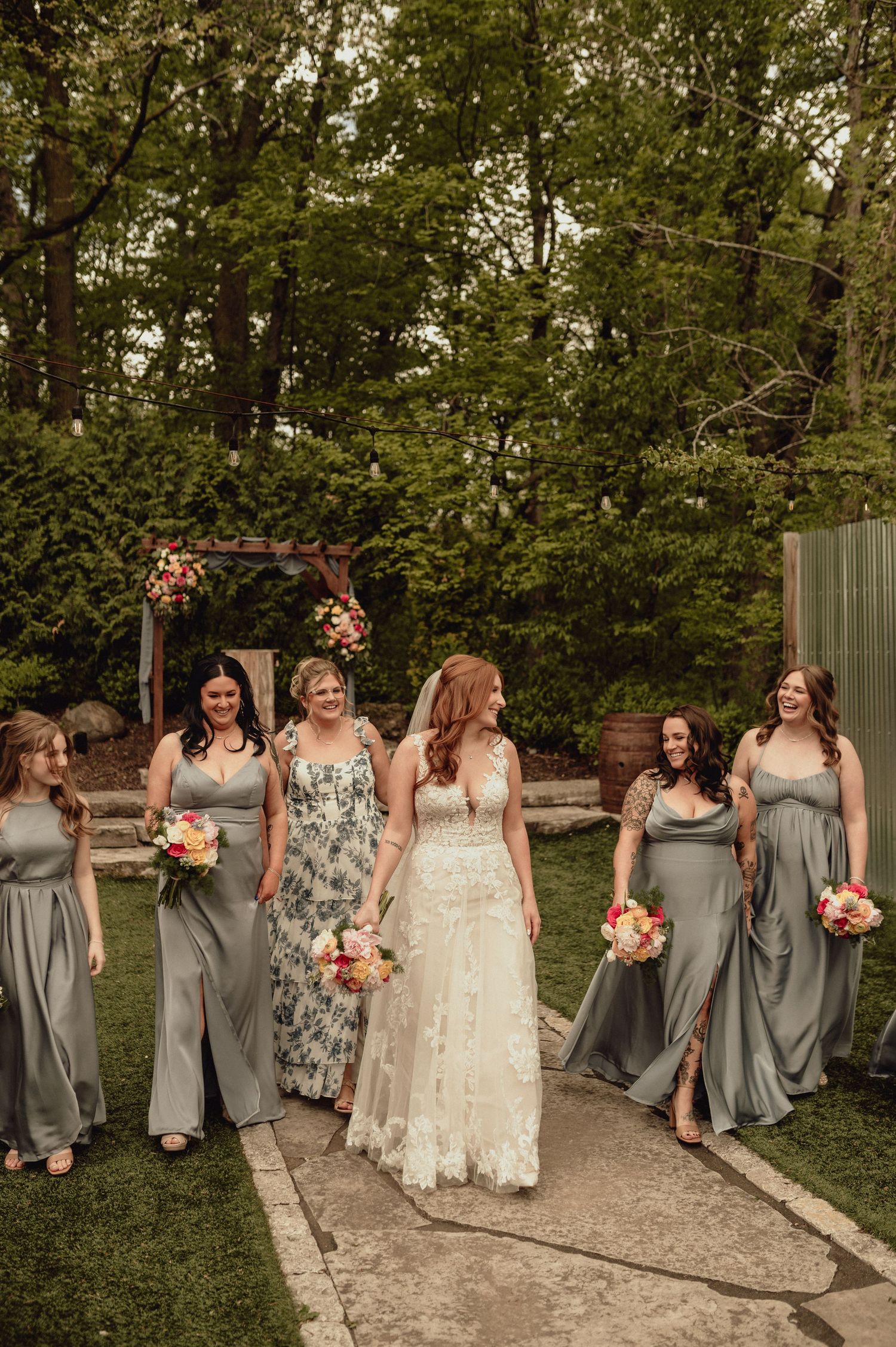 Bridal party in gray dresses and a bride in white walk along a garden path while holding colorful bouquets.