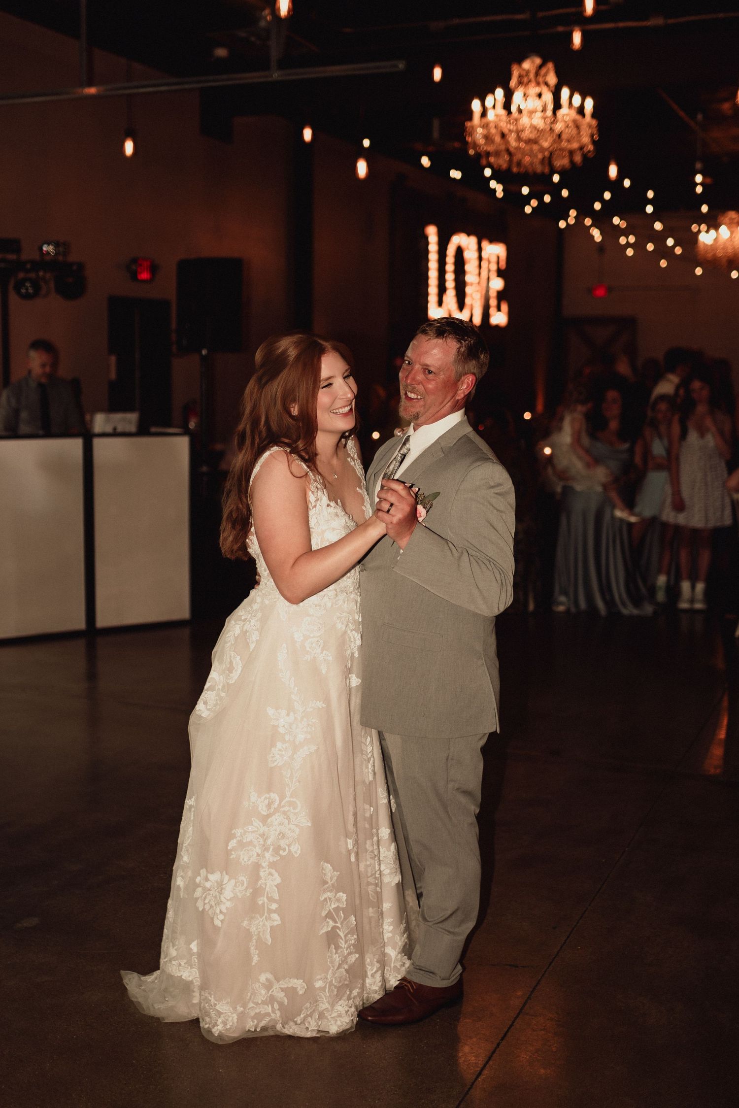 A couple shares their first dance at a wedding reception under a crystal chandelier with illuminated LOVE sign in background.