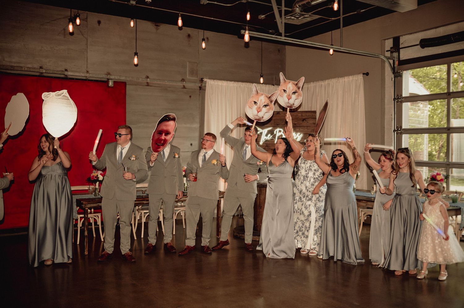 Wedding party in gray dresses celebrates with raised arms in a rustic indoor venue with string lights.