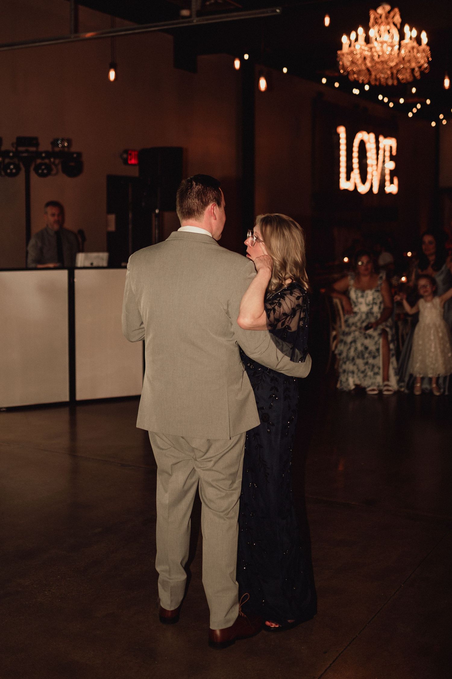 A couple dances together in a dimly lit reception venue with a glowing LOVE sign and chandelier in the background.