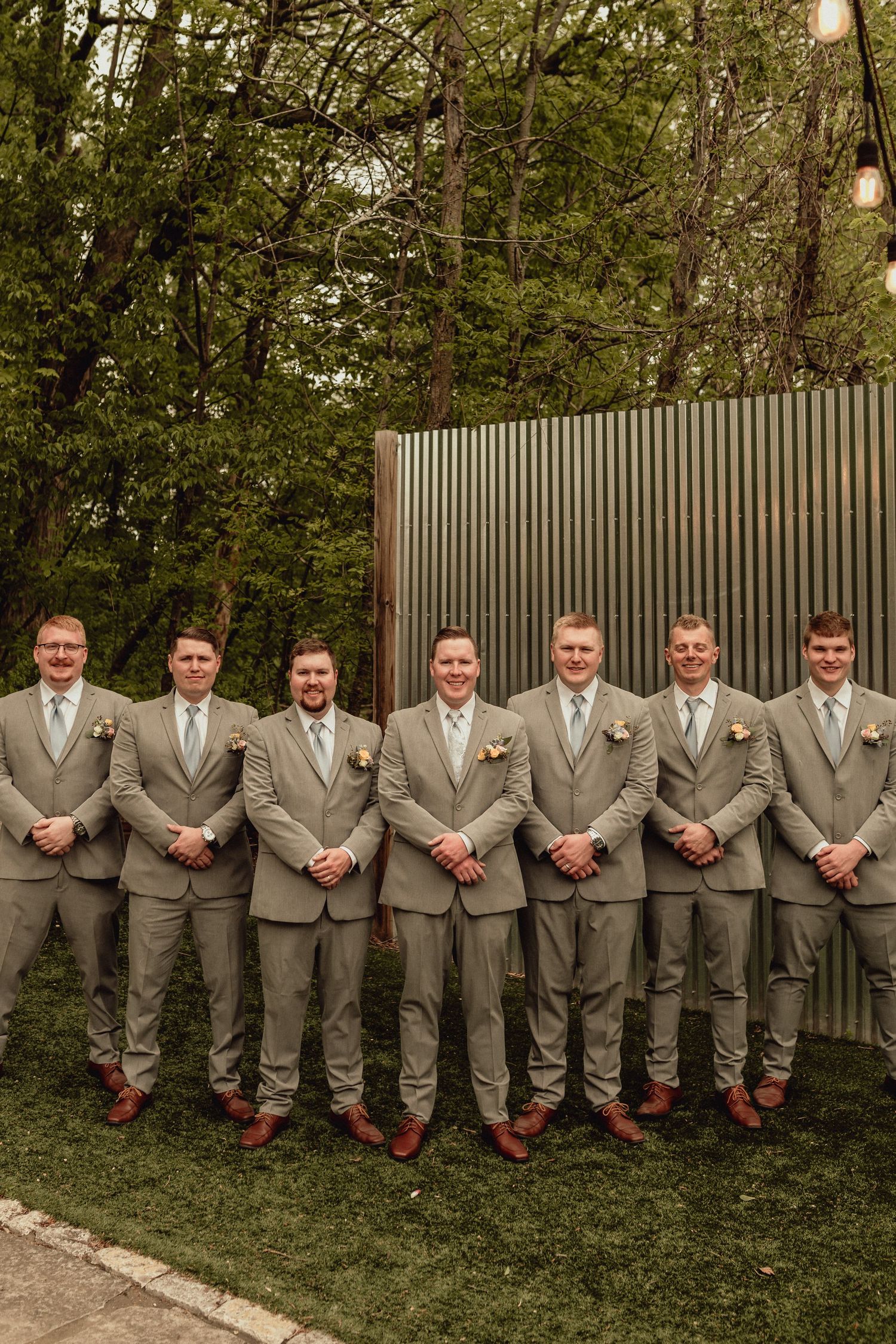 A row of groomsmen in matching gray suits and red socks pose together against a garden fence at an outdoor wedding.