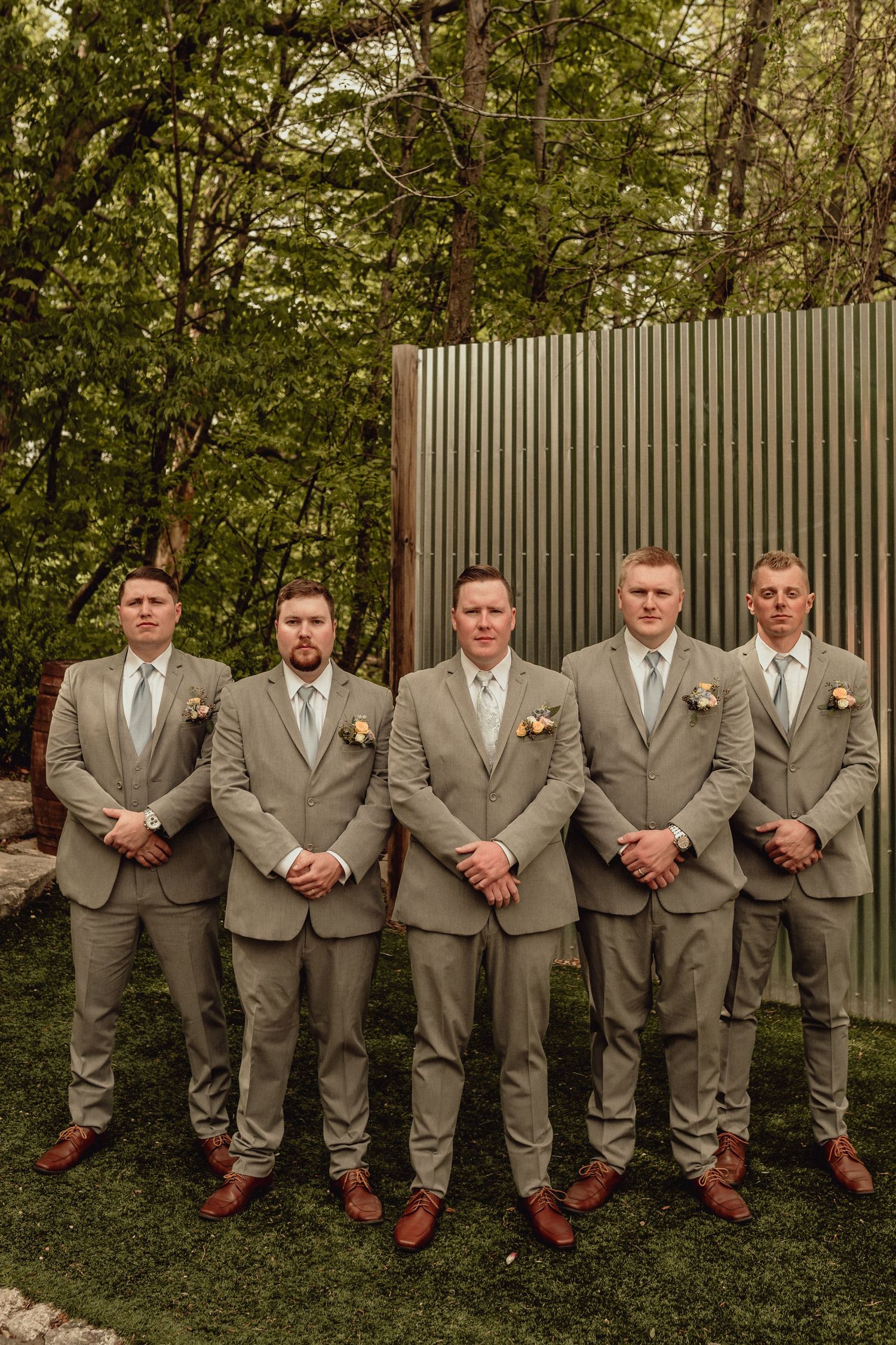 Groomsmen in matching gray suits and white ties lean against wooden fence in outdoor garden setting.