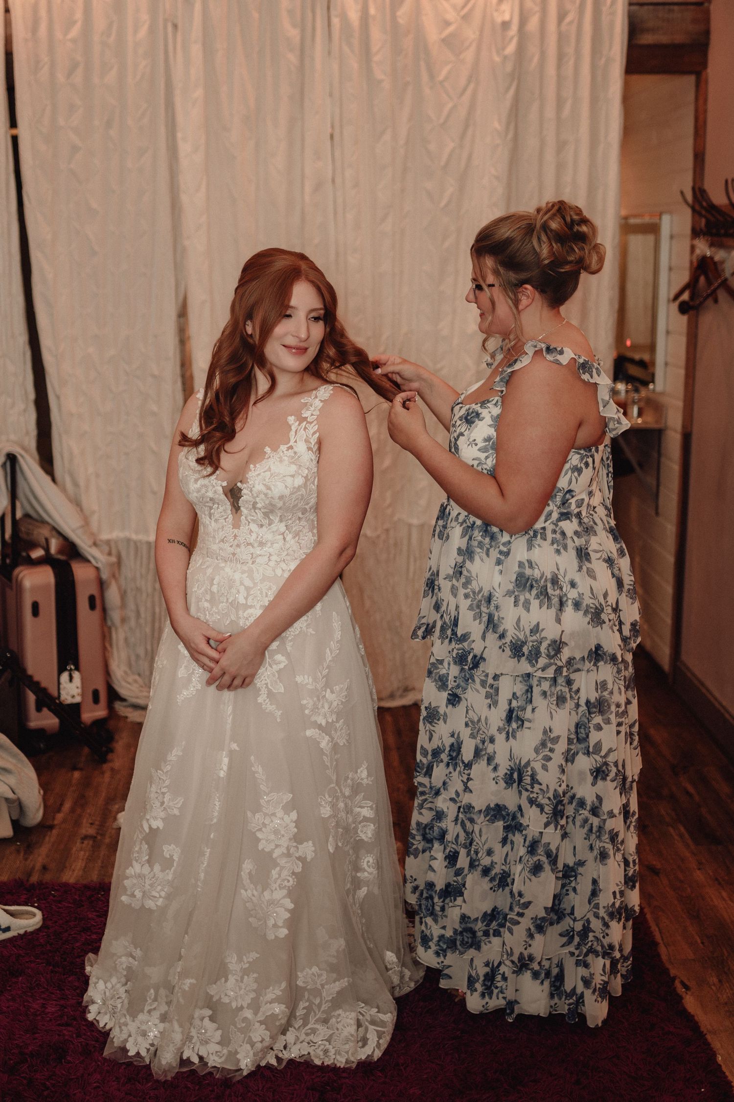 A bride in a white lace gown has her hair styled by someone wearing a blue floral dress in a warmly lit room.