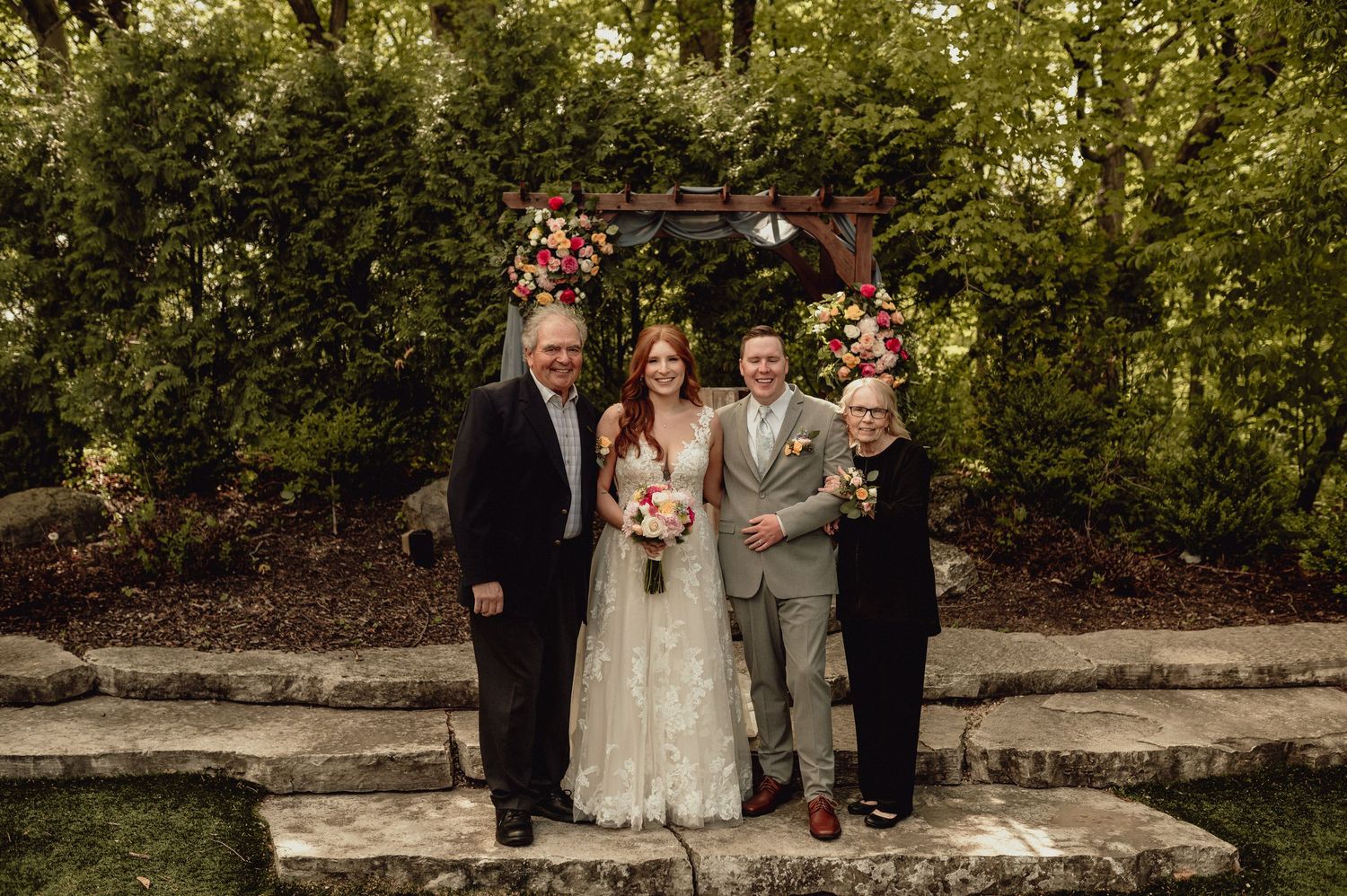 Wedding party poses together on stone steps surrounded by lush greenery in an outdoor garden setting.