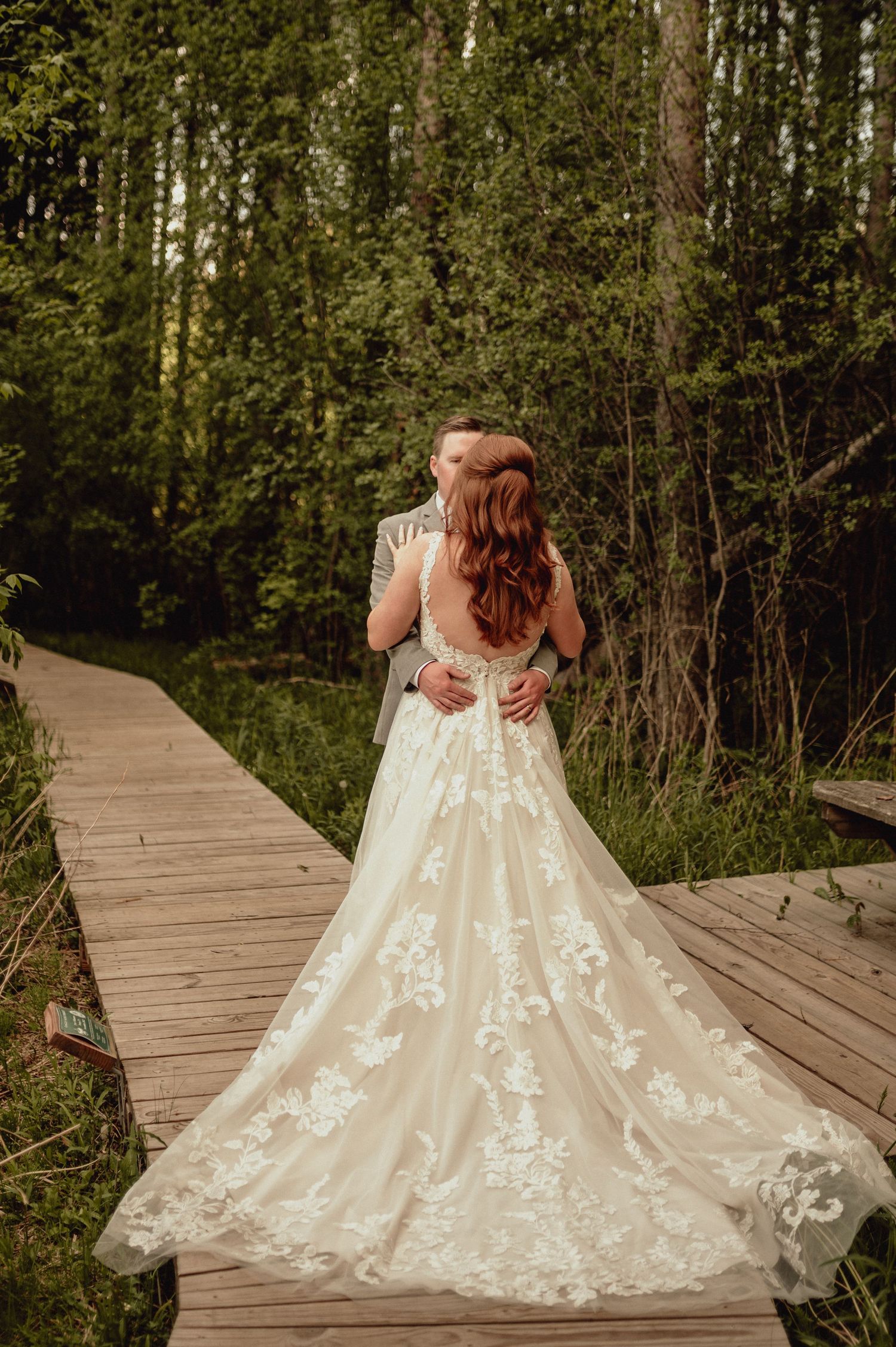 A romantic wedding photo on a wooden boardwalk shows a beautiful lace dress with intricate floral details and flowing train.