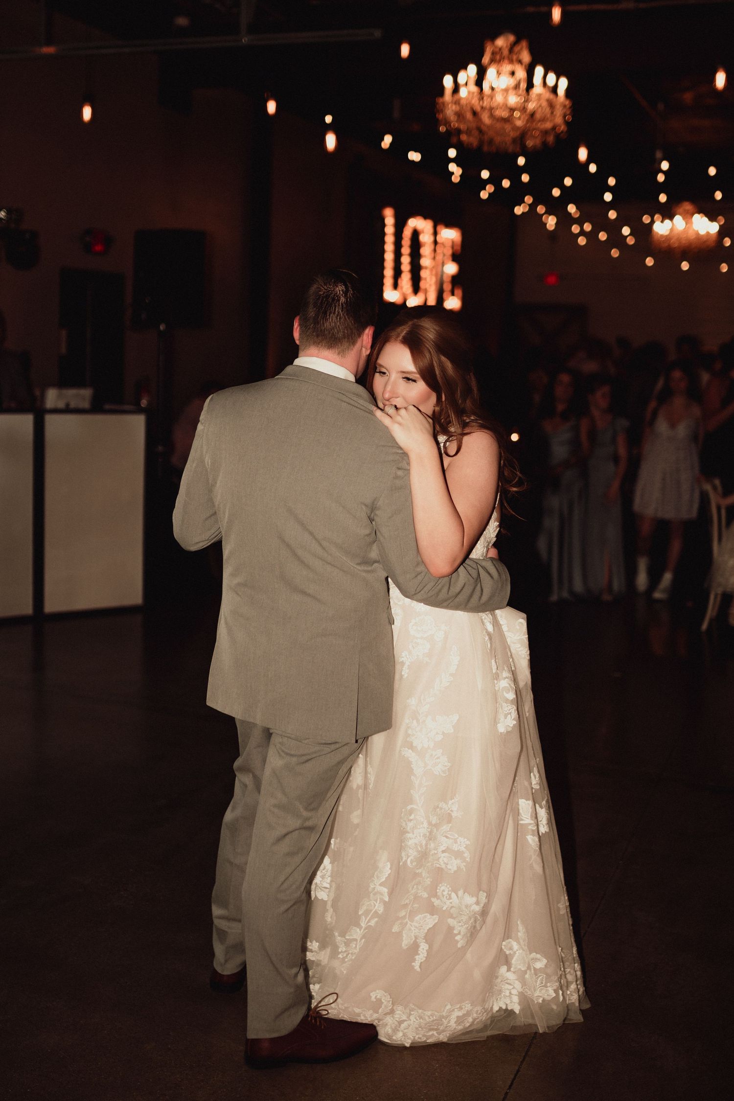 A couple dances together at a dimly lit wedding reception with string lights and chandeliers overhead.
