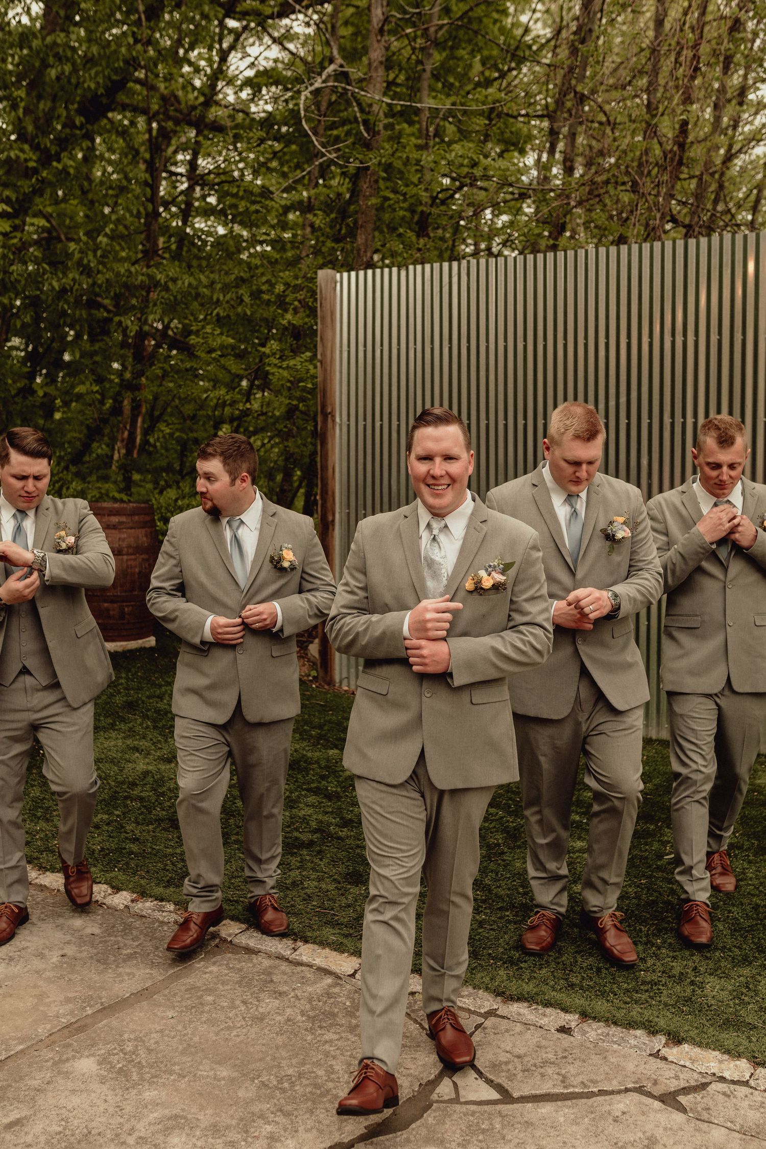 Groomsmen in matching tan suits and brown shoes stand together outdoors against a metal wall and greenery.
