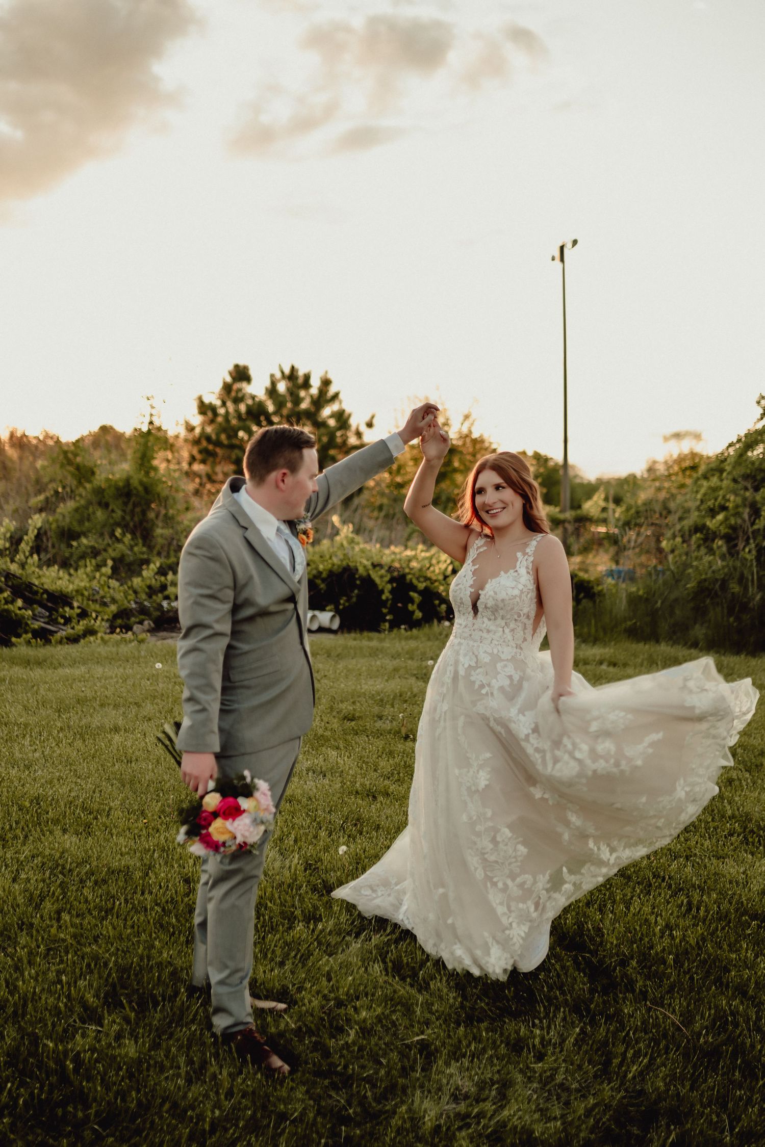 A couple in wedding attire dances together on a grassy lawn at sunset, with the bride's dress flowing in the breeze.