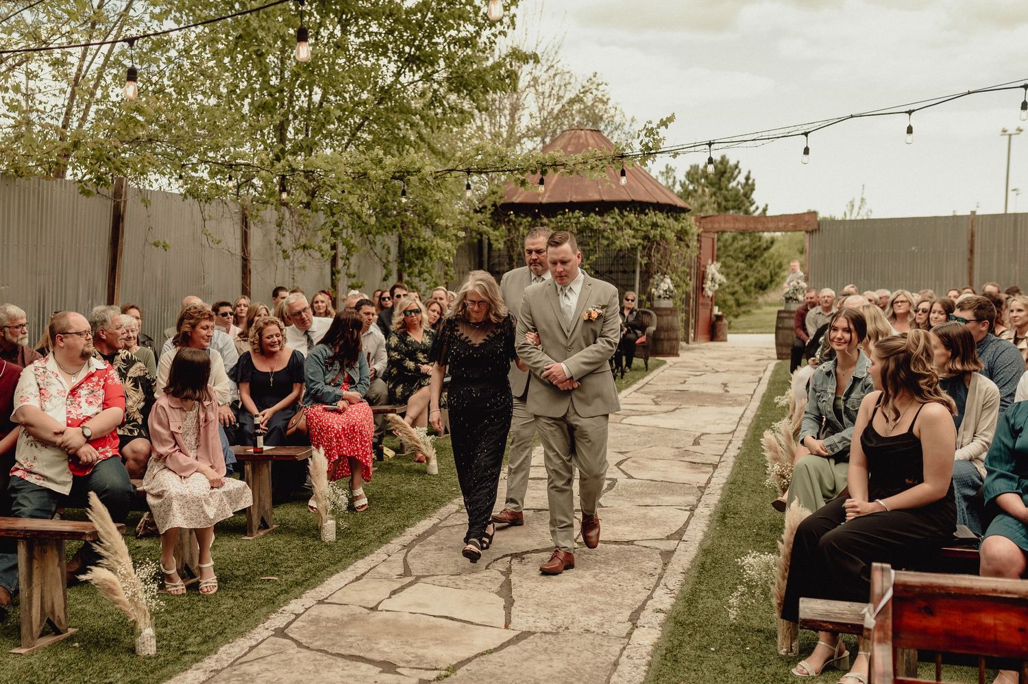 A wedding ceremony takes place outdoors with guests seated on wooden benches under string lights and trees.