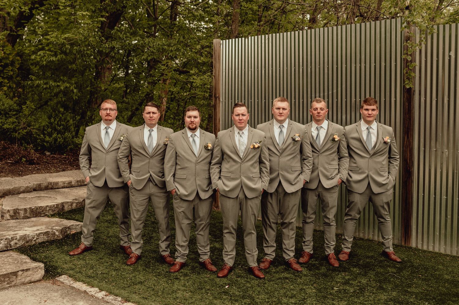 Wedding party in matching gray suits and brown shoes pose together against a wooden fence backdrop.
