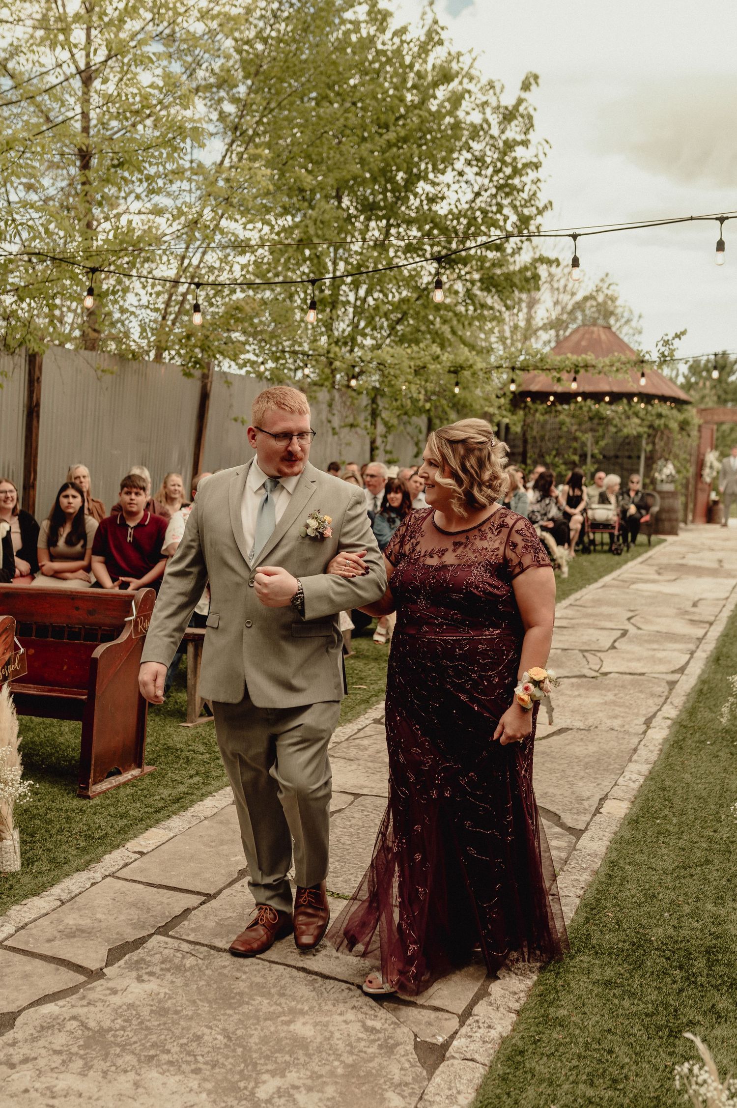 Two people dance together at an outdoor wedding reception on a stone pathway surrounded by trees and string lights.