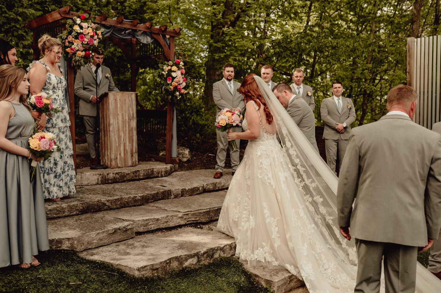 Outdoor wedding ceremony under floral arch with wedding party in sage green and gray attire standing on stone steps.