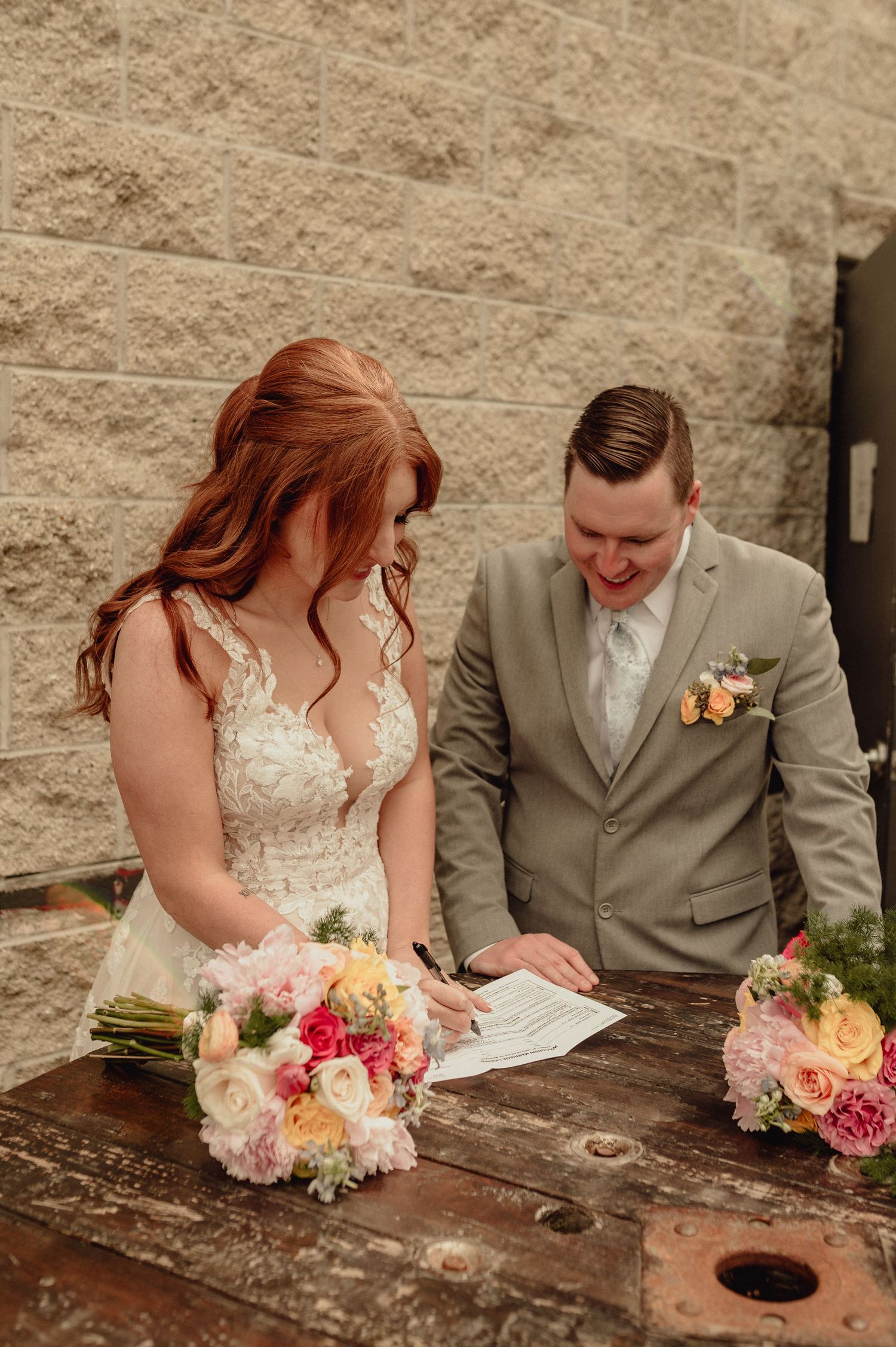 Newlyweds signing marriage documents together at rustic wooden table with colorful floral bouquet.