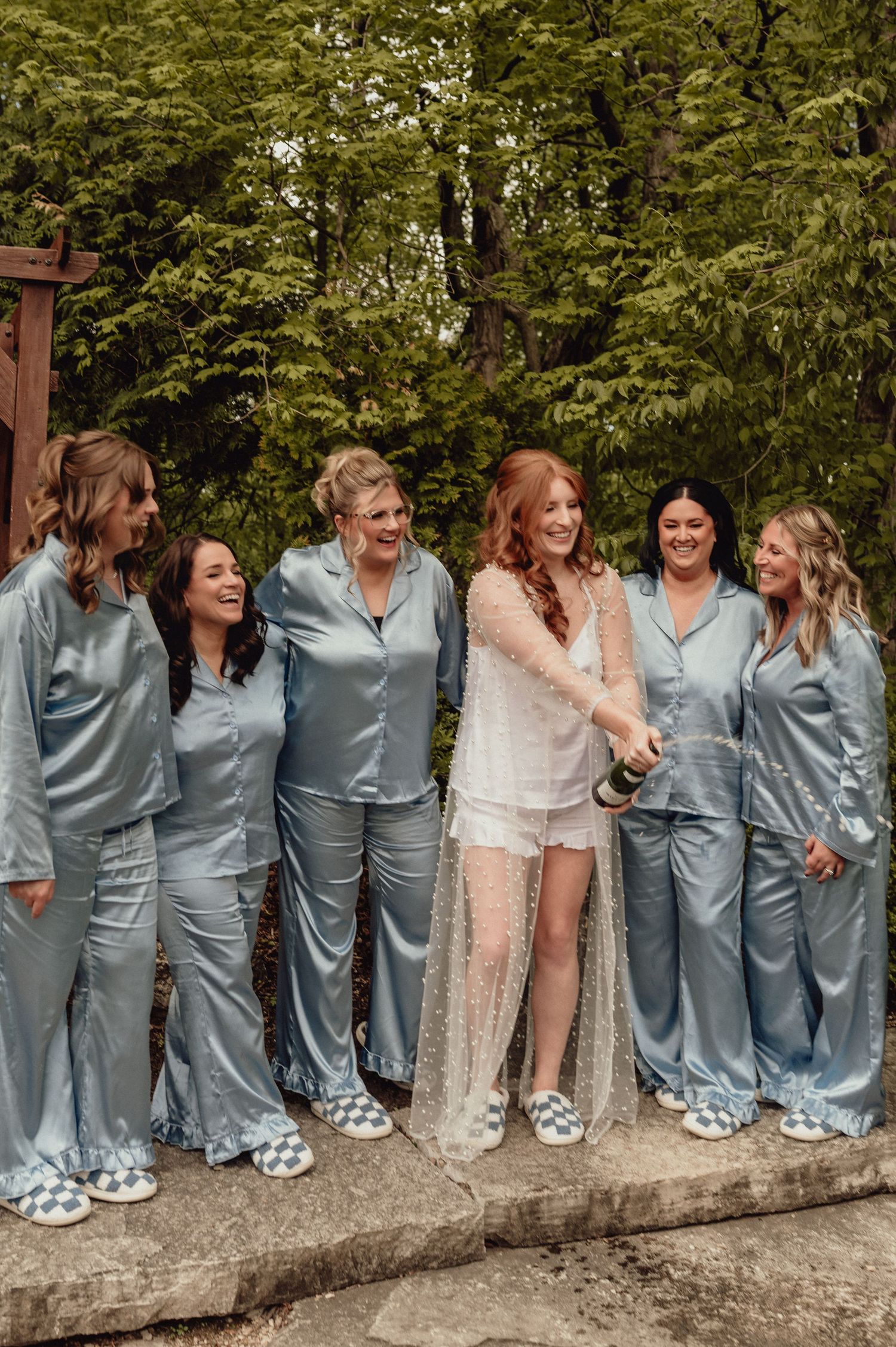 Wedding party in matching blue outfits and checkered slippers pose together on a stone wall in a garden setting.