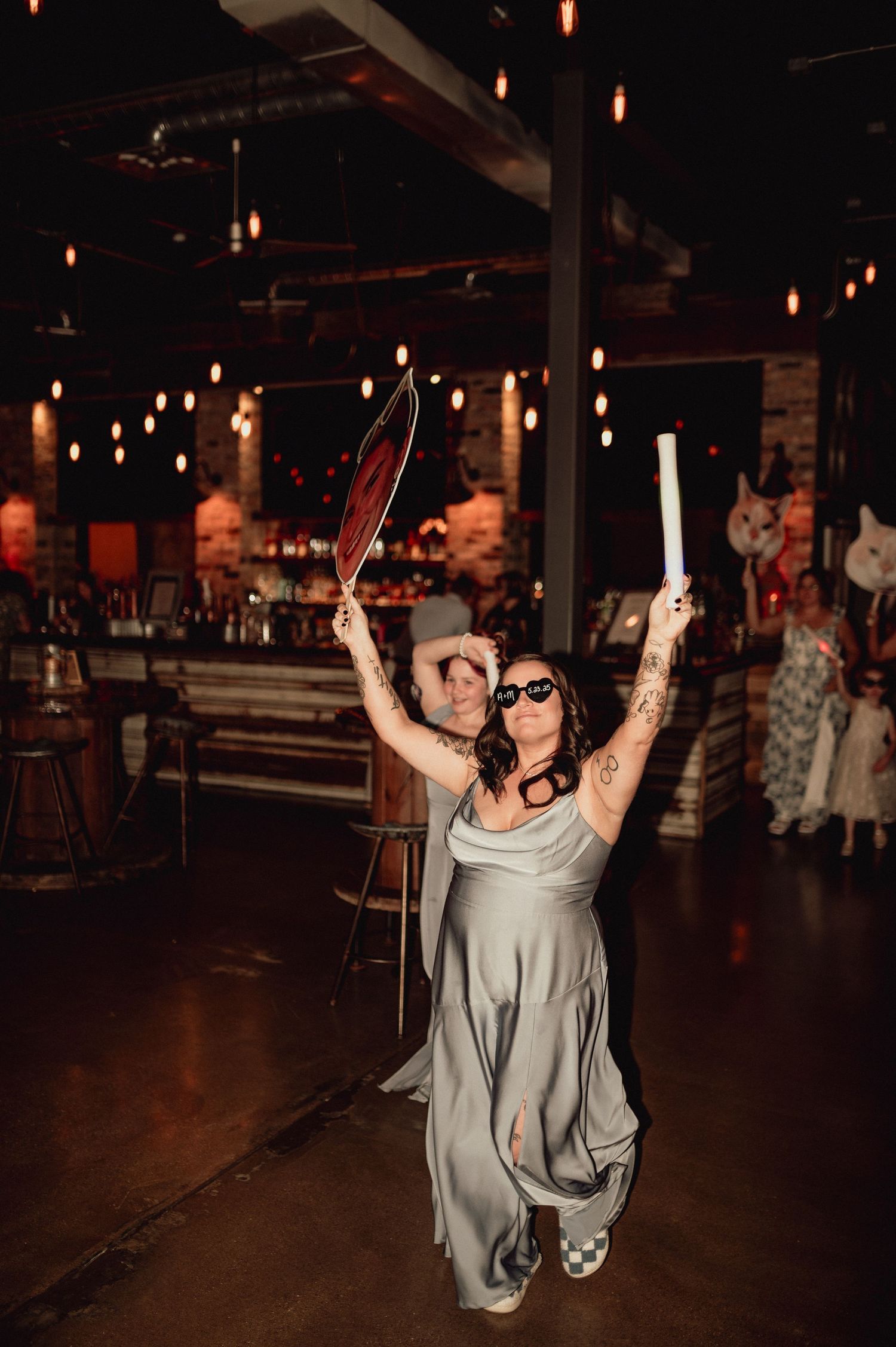 A person in a gray dress and sunglasses dances enthusiastically on a dark dance floor with overhead lighting.