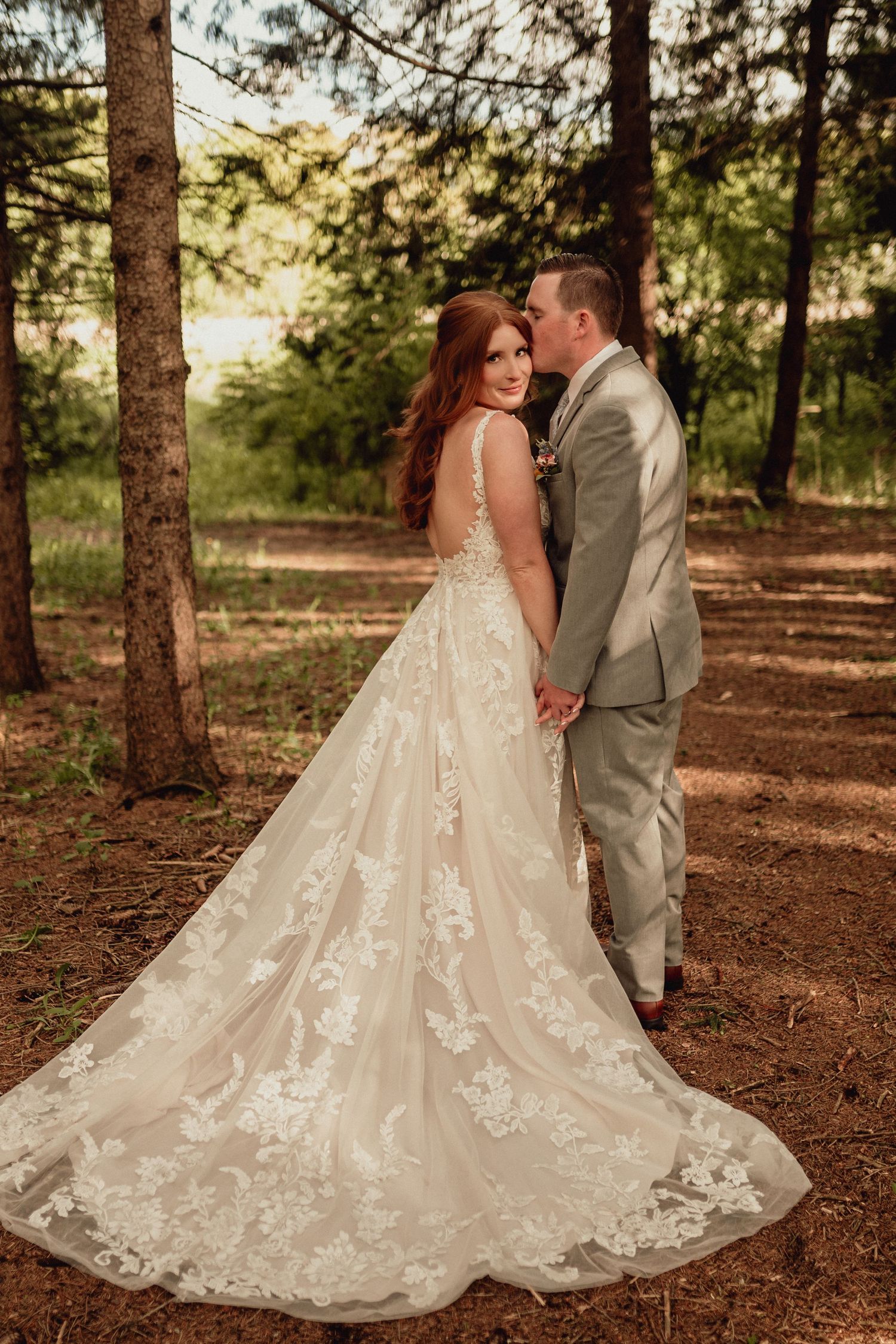 A couple in wedding attire embraces in a sunlit forest setting with the bride wearing a flowing lace gown with a dramatic train.