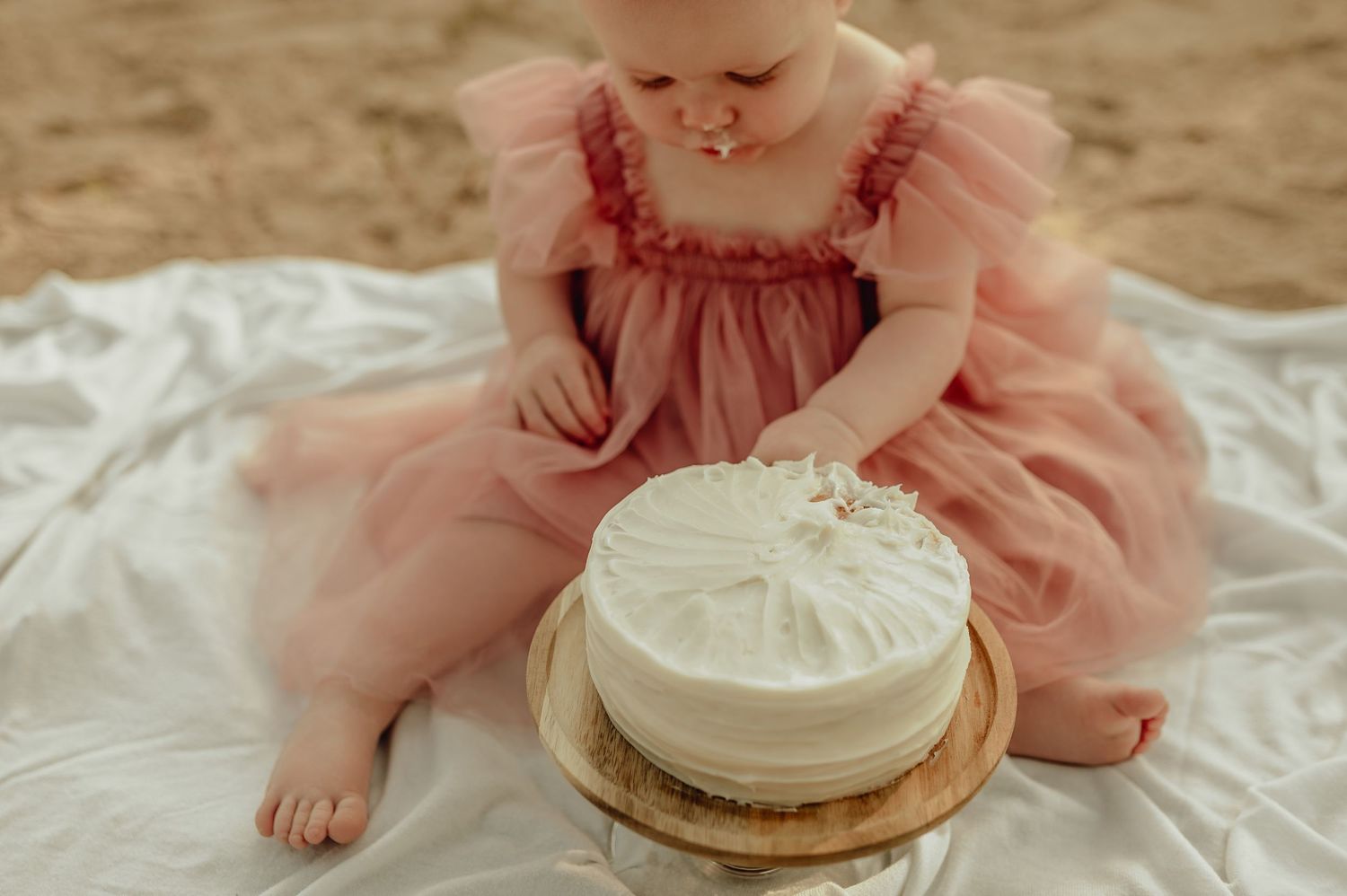 Baby wearing pink dress reaching for white cake during outdoor photo session.