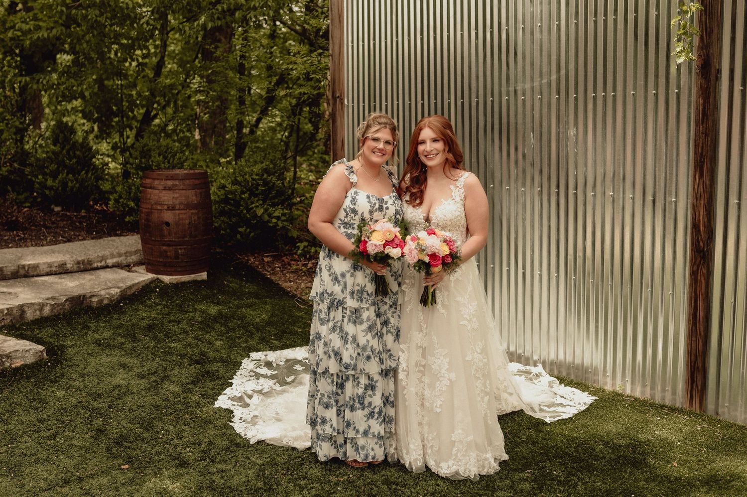 Wedding party members in floral gray dresses pose with bride in white lace gown against rustic metal wall backdrop.