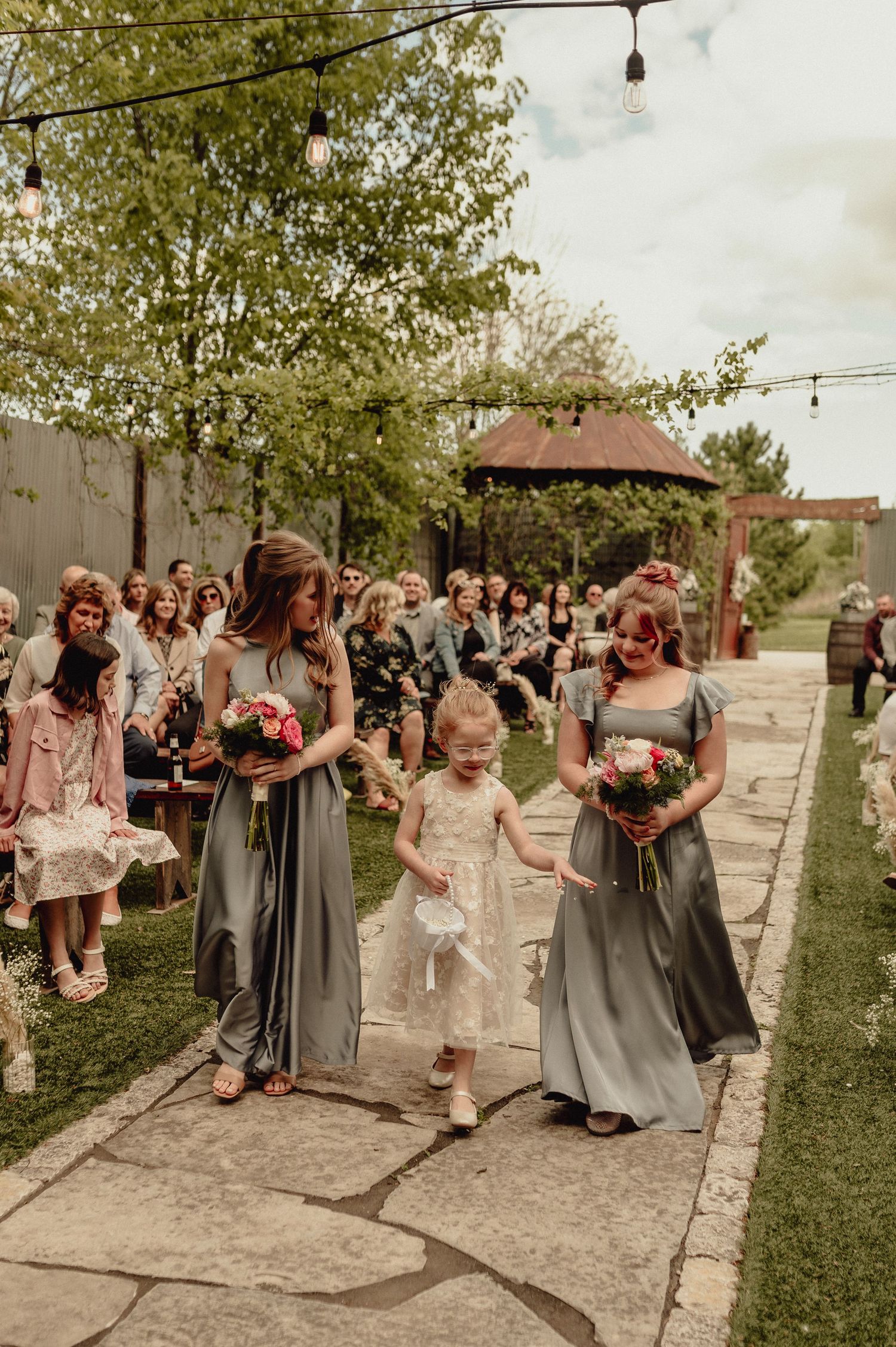 Wedding party members dance and celebrate outdoors on a stone path lined with trees during a summer celebration.