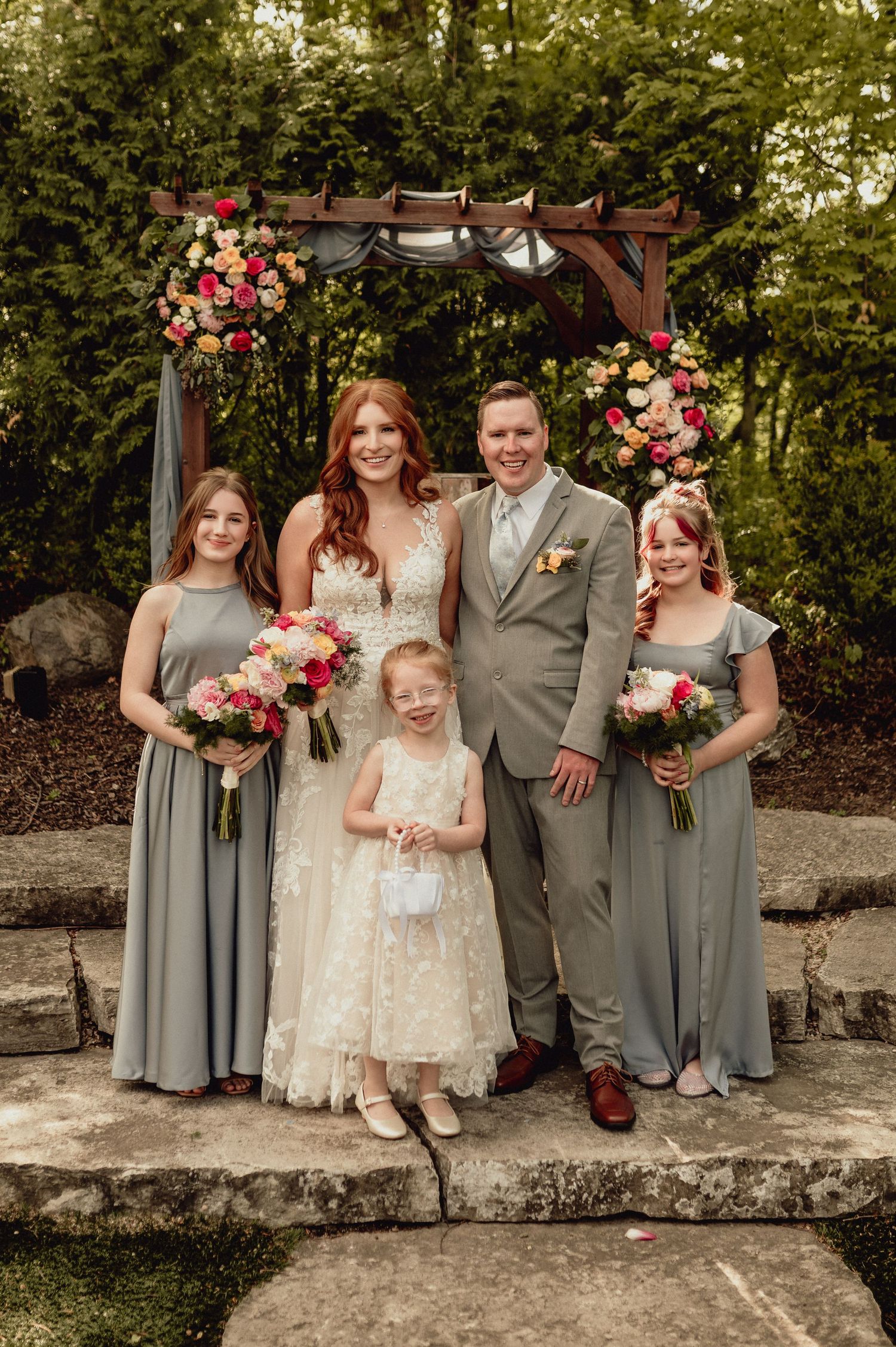 Wedding party in gray and white attire poses together outdoors with pink floral bouquets and decorations.