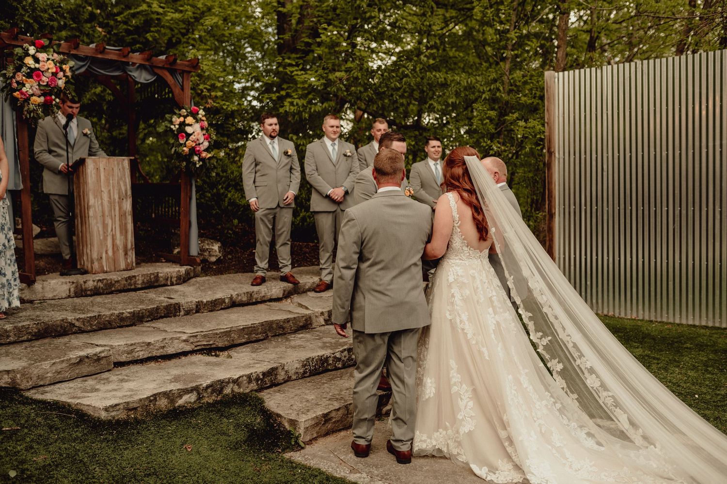 Wedding party poses in garden setting with bride in flowing white gown and groomsmen in light gray suits.
