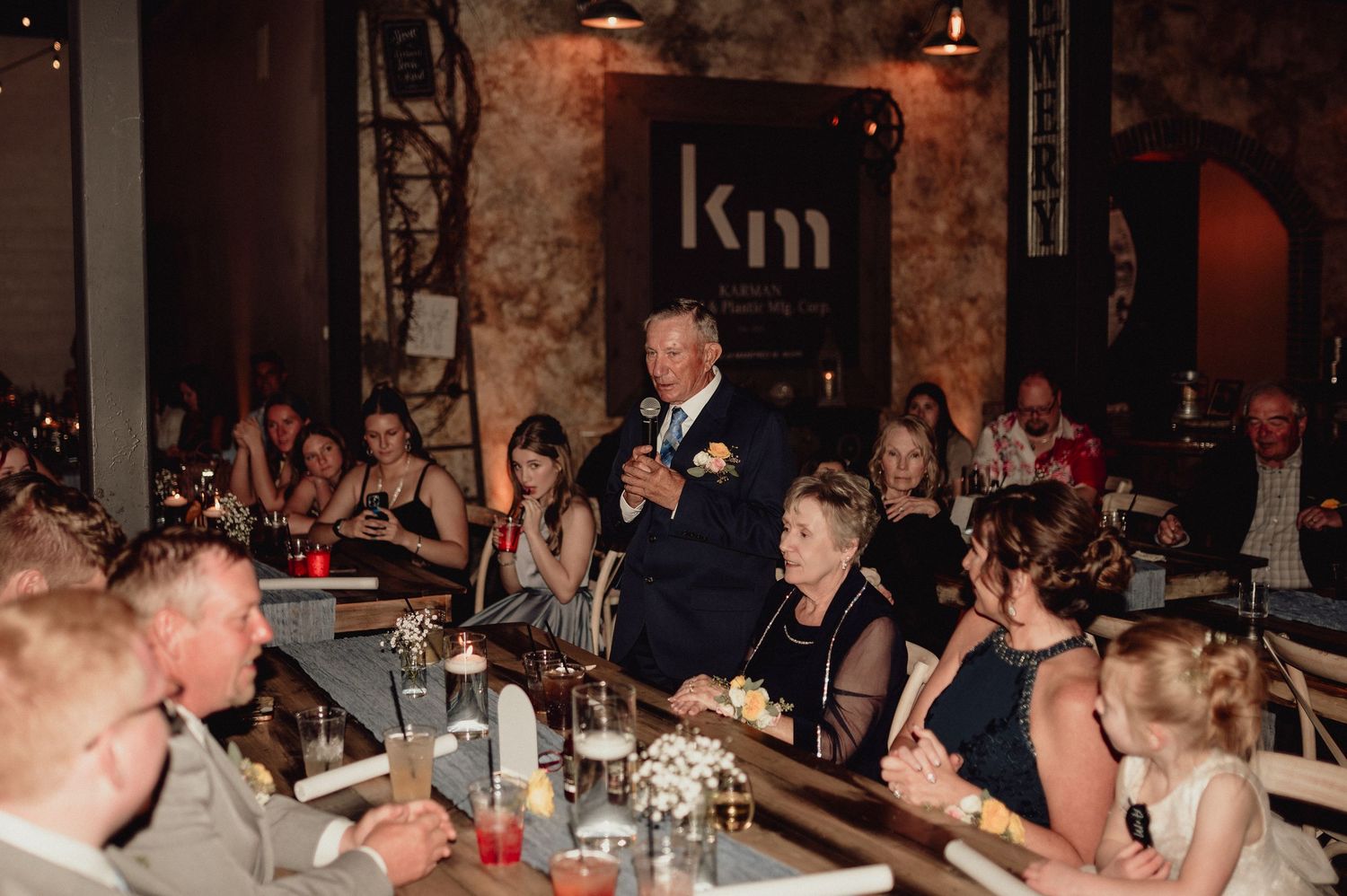 A lively wedding reception with guests seated at tables in a dimly lit rustic venue with stone walls.