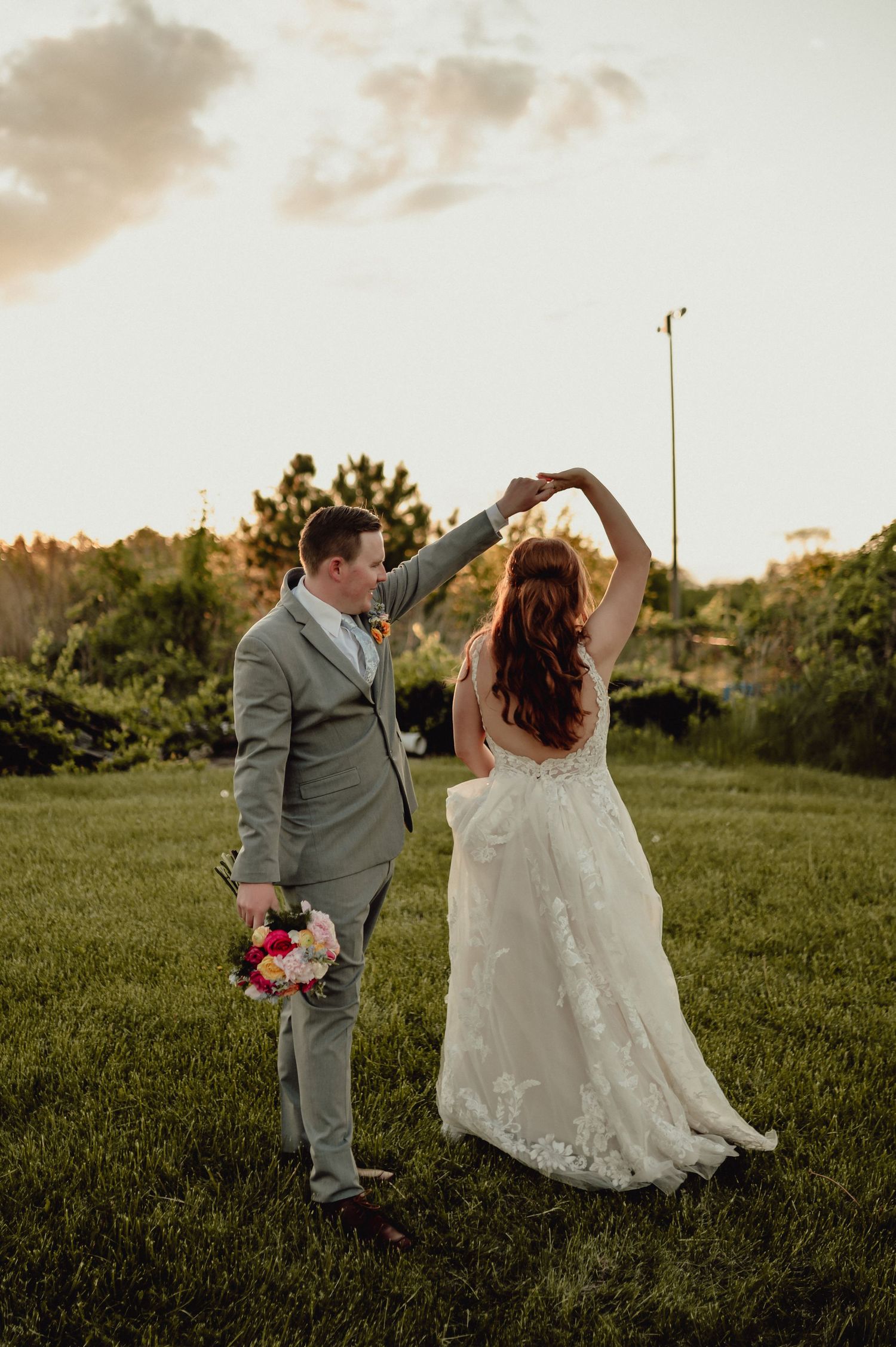 A bride and groom dance together in a grassy field at sunset, creating a romantic silhouette against the warm sky.