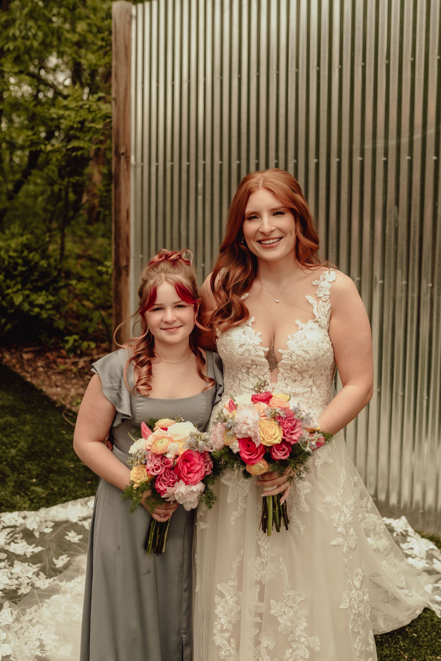 A bride in a lace wedding gown poses with a bridesmaid in grey holding vibrant pink and white floral bouquets.