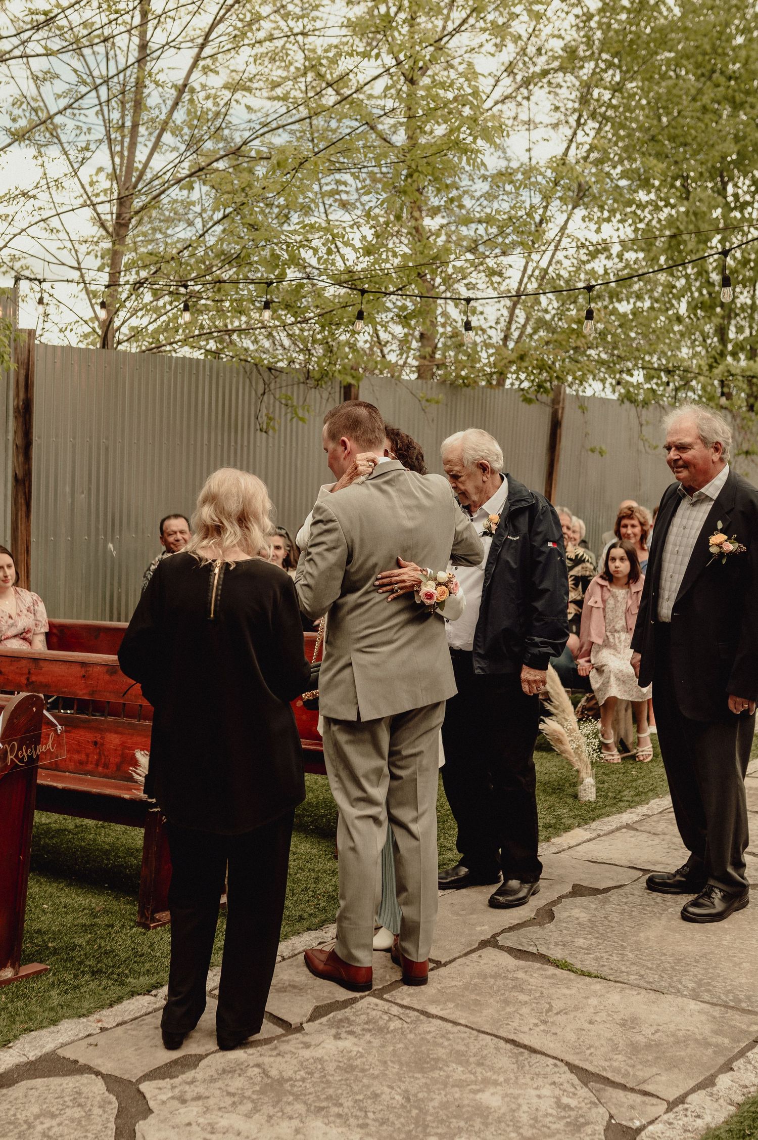 Wedding guests gather outdoors in formal attire near a red bench and wooden fence during a daytime celebration.