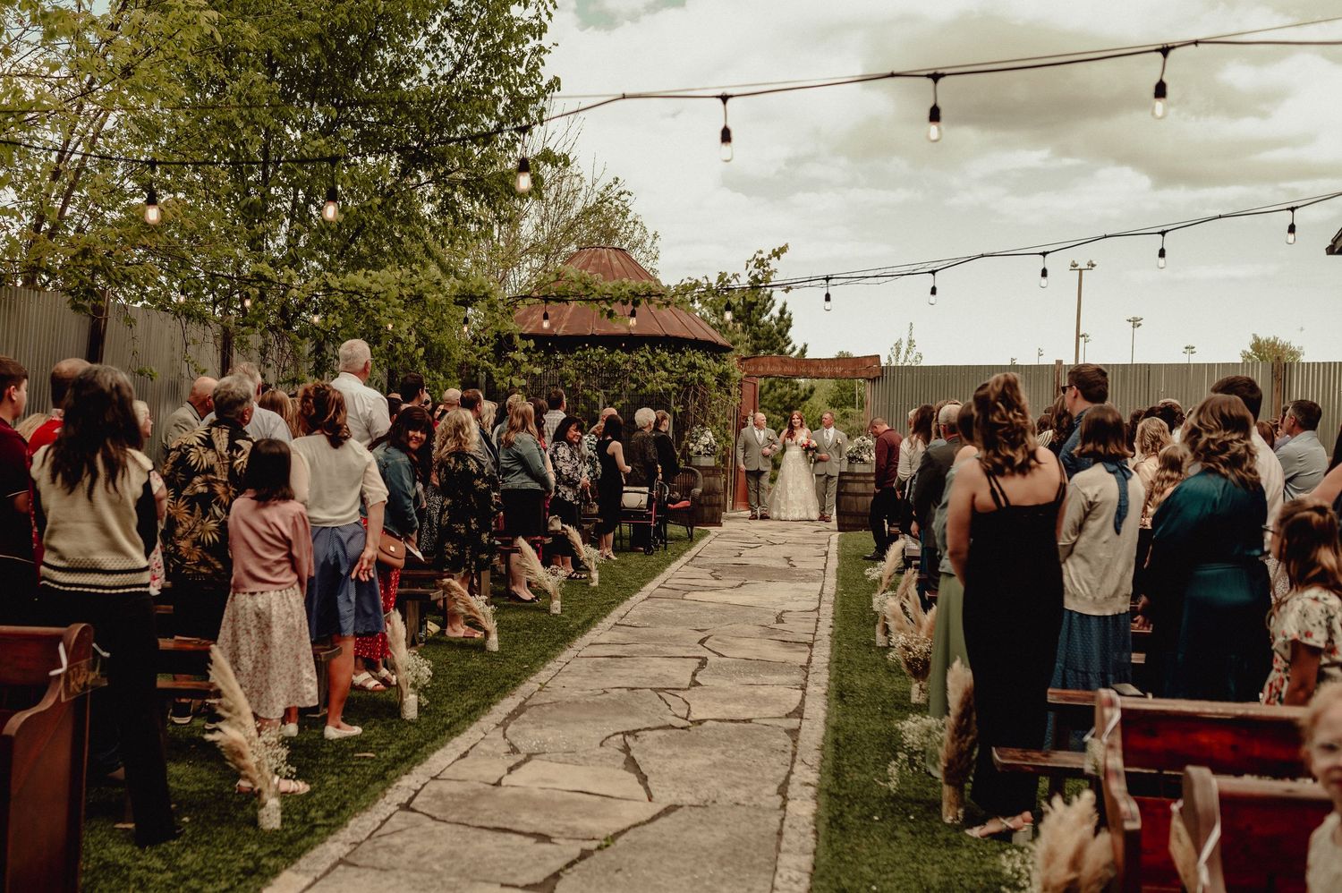 String lights illuminate an outdoor wedding ceremony with guests gathered under trees at sunset.